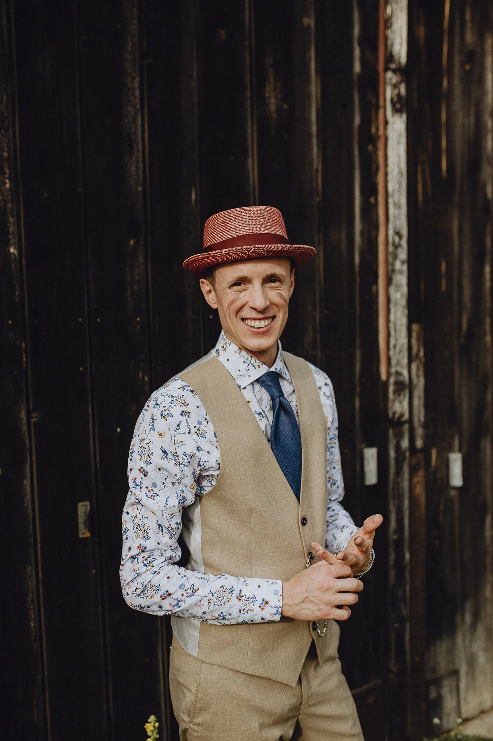 Ein locker gekleideter Bräutigam trägt einen Hut, ein kariertes Hemd, eine Weste und eine Krawatte, lächelt und steht vor einer dunklen Holzwand.