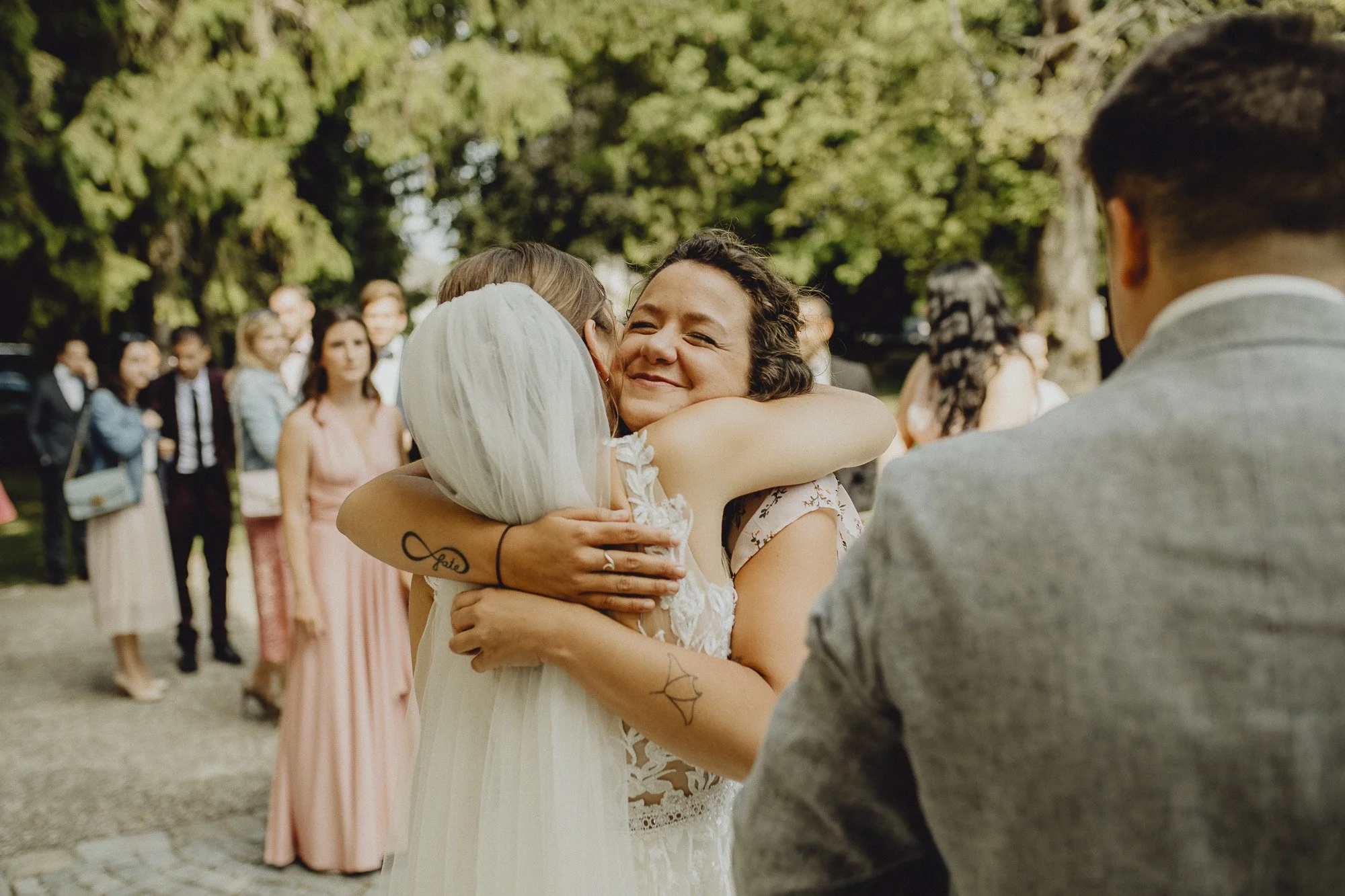 Eine Frau mit kurzen dunklen Haaren umarmt eine Person im weißen Brautkleid bei einer Hochzeit im Freien, umgeben von Gästen, die im Hintergrund stehen.