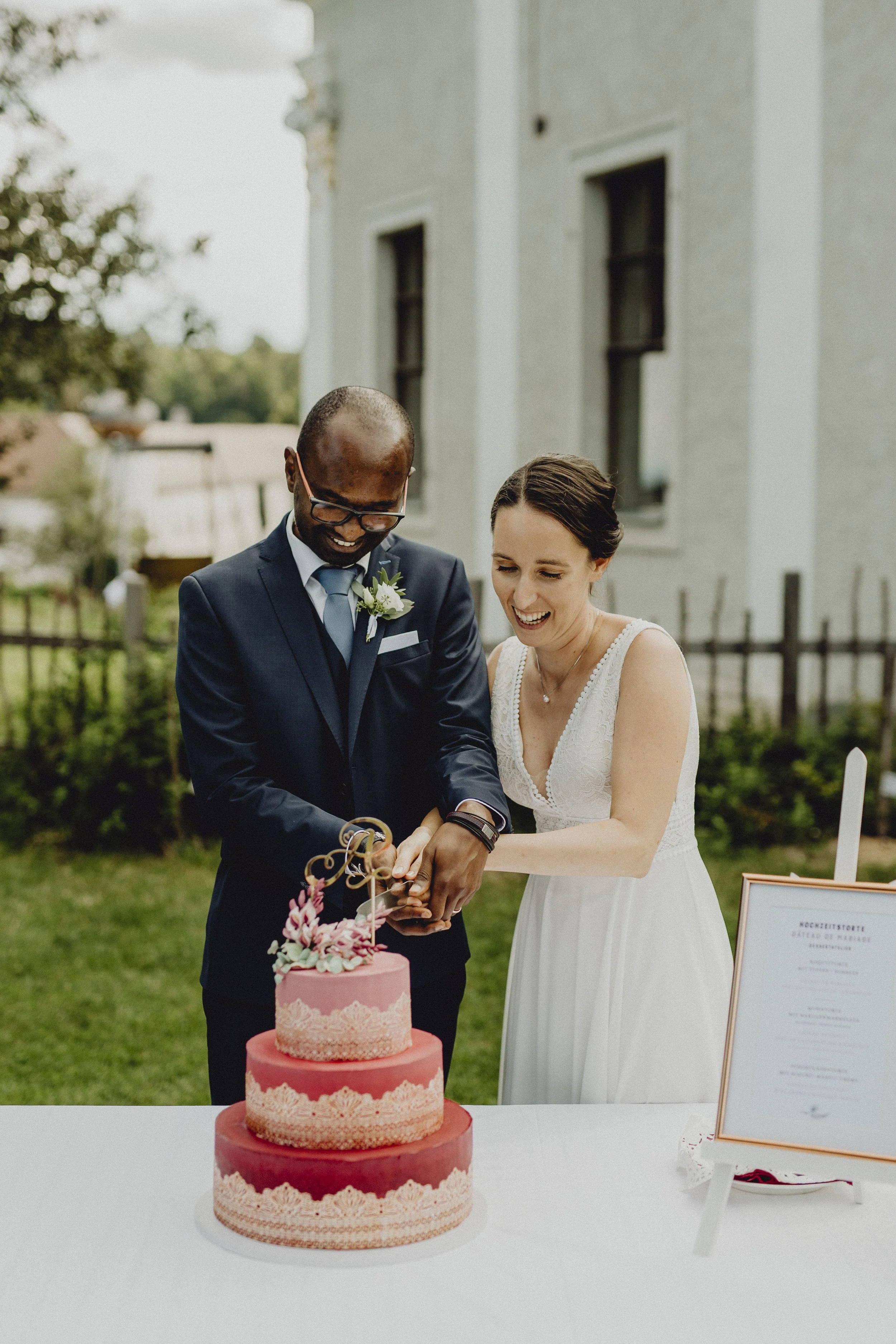 Das Brautpaar schneidet gemeinsam die Hochzeitstorte, beide lachen fröhlich, draußen bei Sonnenschein, im Hintergrund ein historisches Gebäude.
