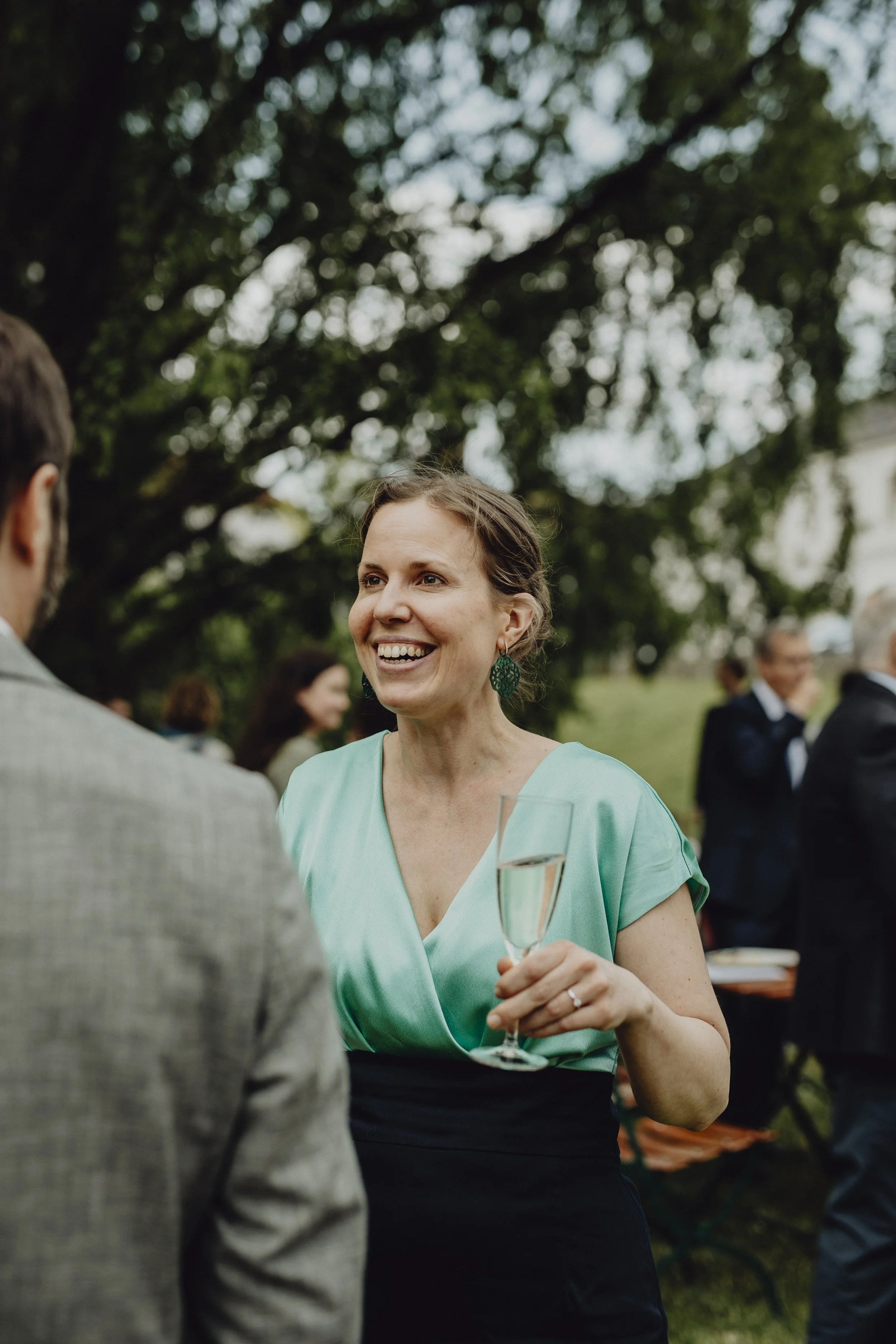 Eine Frau in einem türkisen Kleid hält ein Champagnerglas bei einer einer Hochzeitsfeier, im Hintergrund sind weitere Gäste.