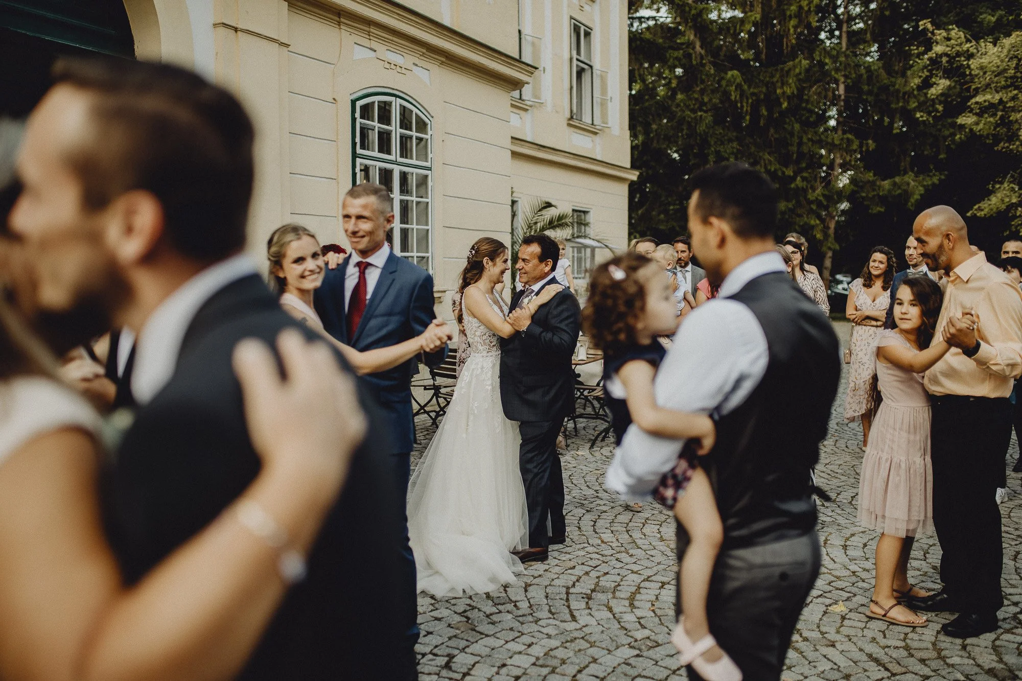 Tanzende Hochzeitsgäste im Außenbereich, Braut und Brautvater in der Mitte, umgeben von Familienmitgliedern und Freunden.