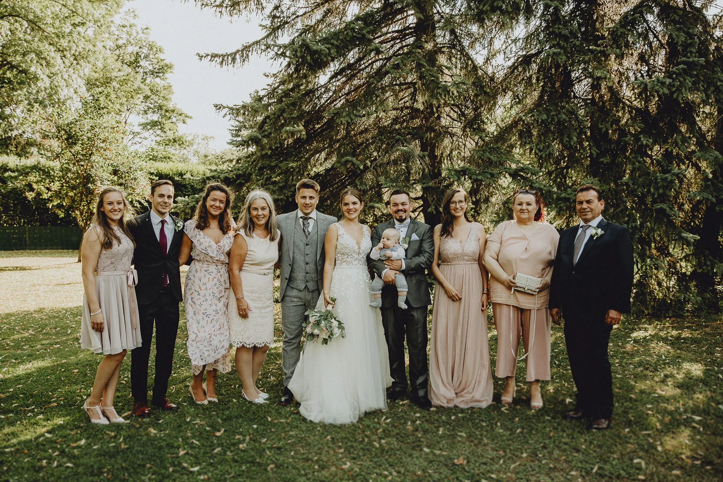 Eine Gruppe von Menschen in festlicher Kleidung steht im Freien vor einem großen Baum. Das Brautpaar steht in der Mitte.