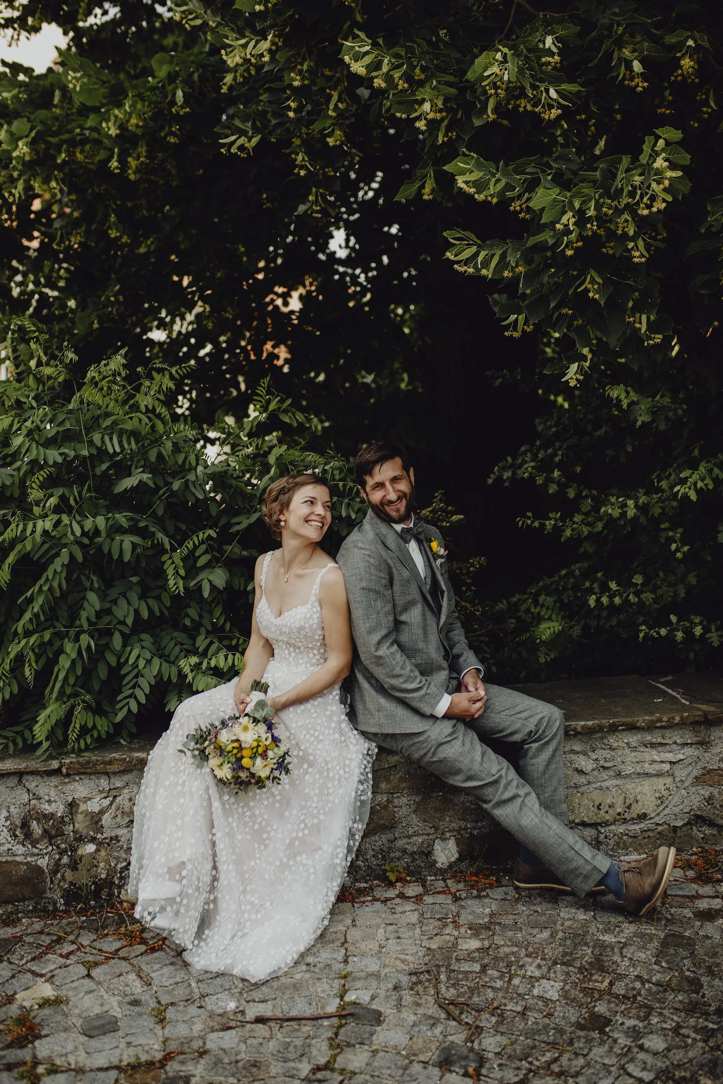 Ein Brautpaar sitzt lachend auf einer Steinmauer, umgeben von grünen Büschen und Bäumen und schauen einander über ihre Schulter an.