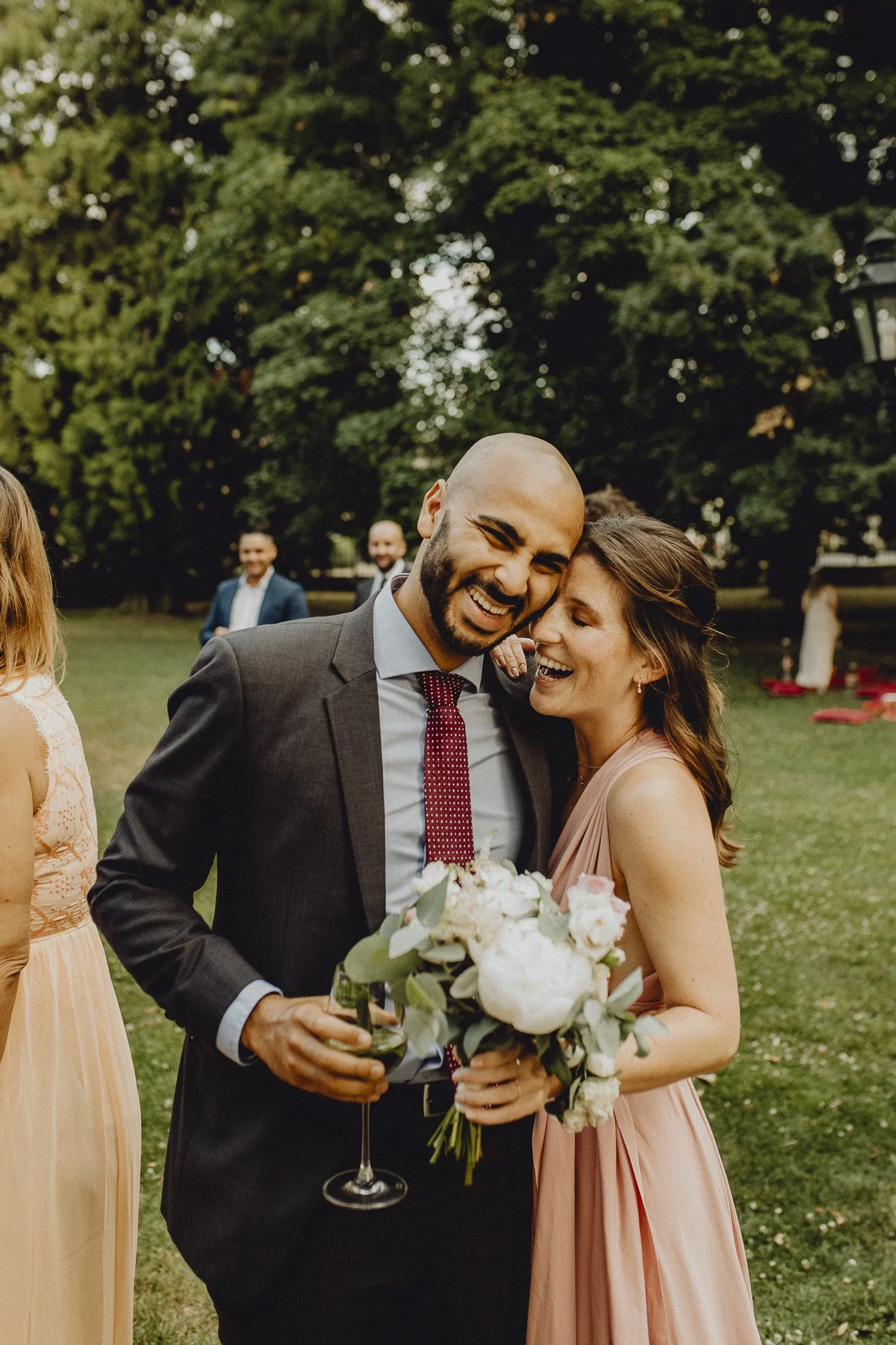 Zwei lachende Menschen bei einer Hochzeitsfeier, eine Frau mit Blumenstrauß und ein Mann mit Glas Wein.