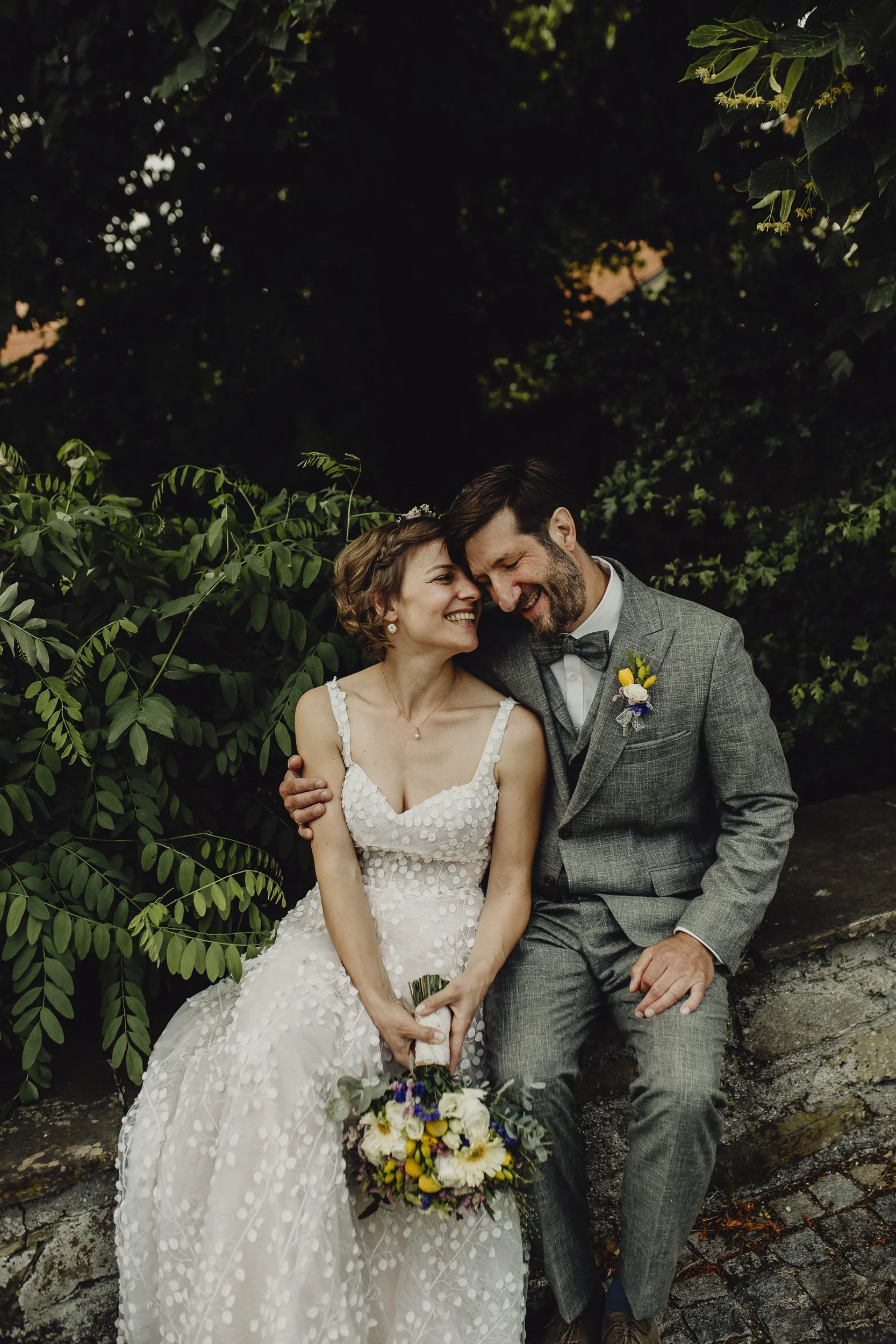 Ein glückliches Brautpaar sitzt draußen an einem vom Laub umgebenen Ort, lächelt sich an und hält einen Blumenstrauß in den Händen.