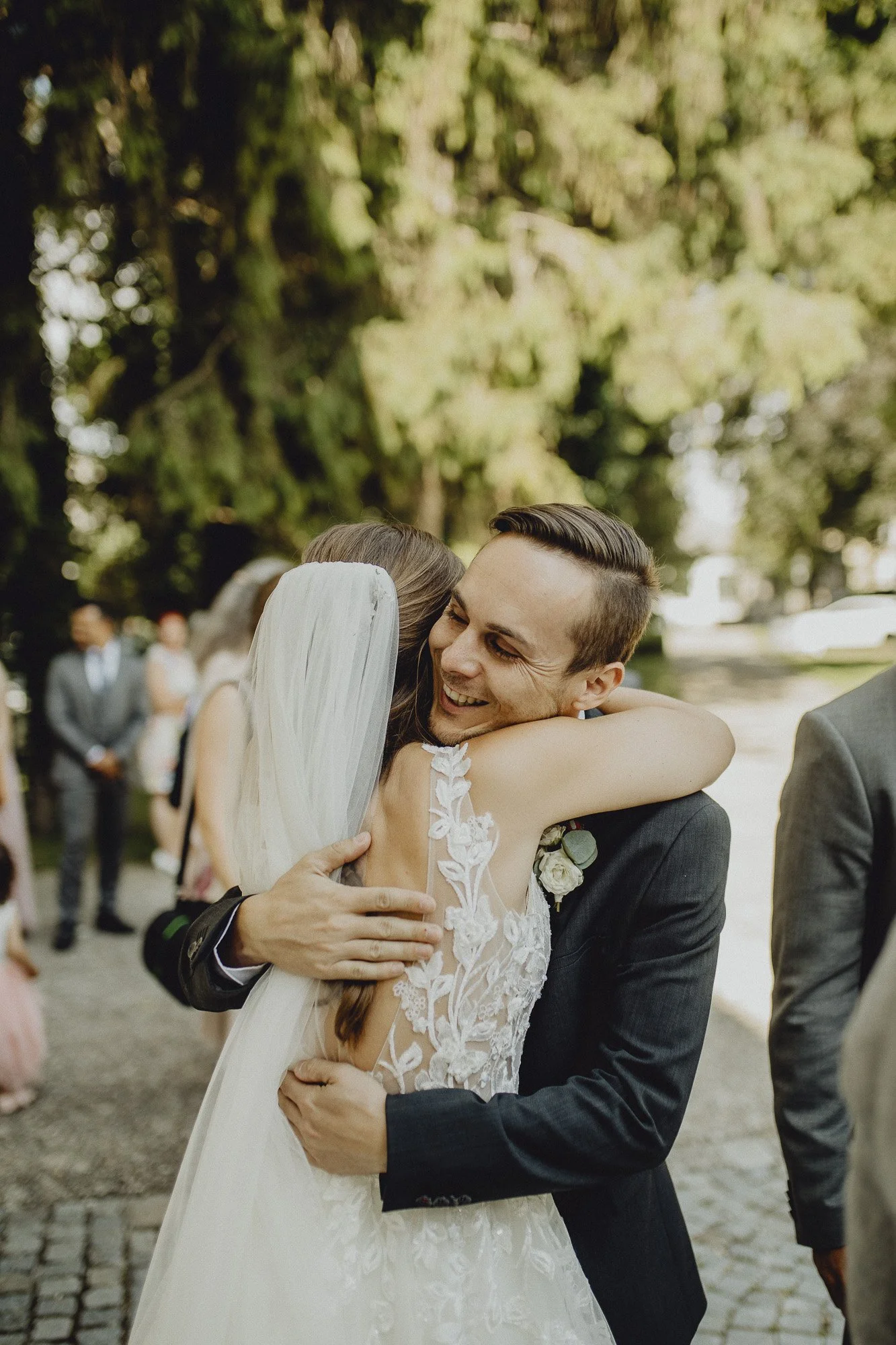 Hochzeitspaar, die sich herzlich umarmen, während Gäste im Hintergrund stehen, draußen unter Bäumen.