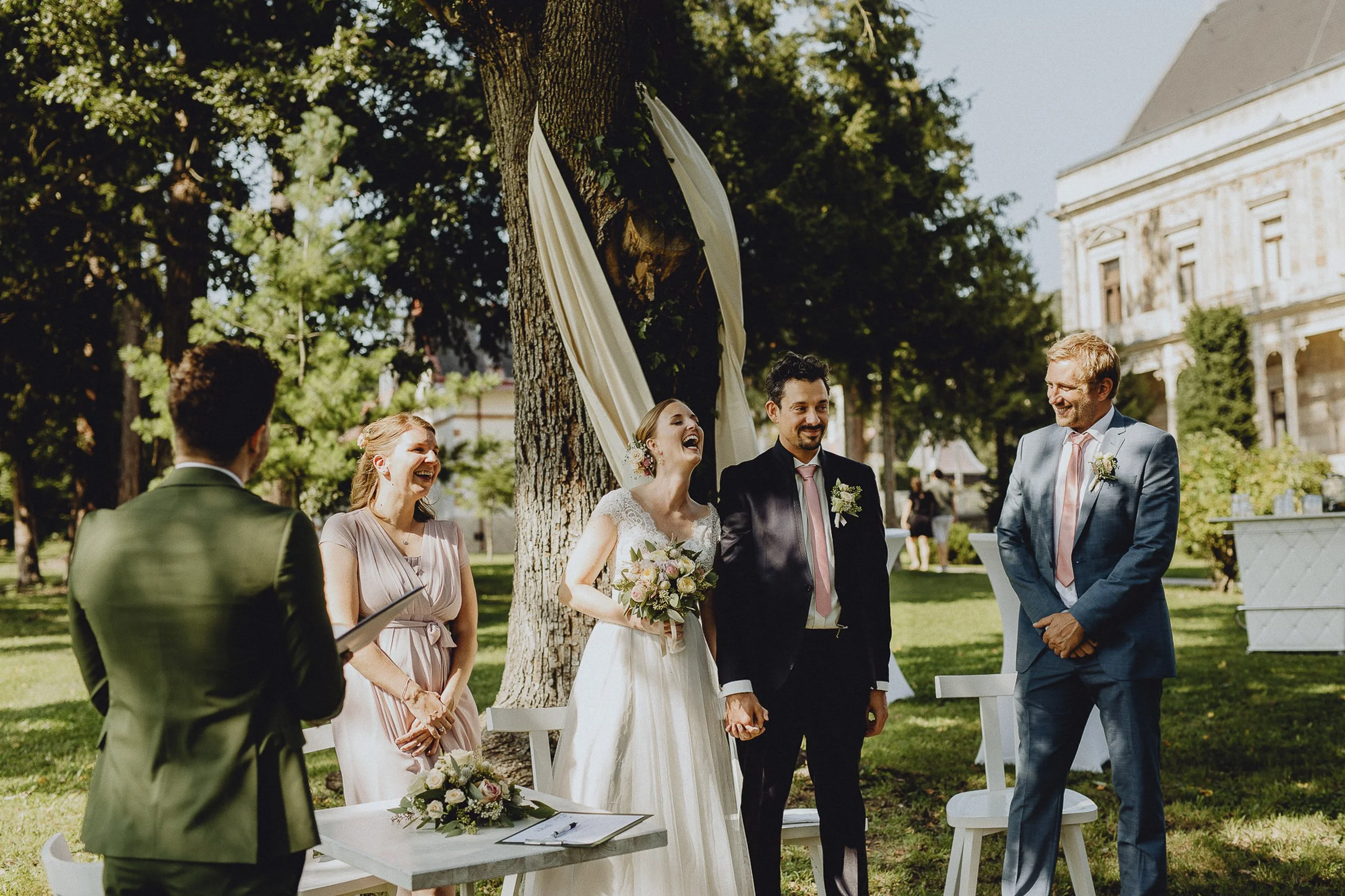 Ein Hochzeitszeremonie im Freien unter einem Baum, bei der ein Paar die Ringe austauscht, umgeben von Freunden und Familie, bei Sonnenlicht.