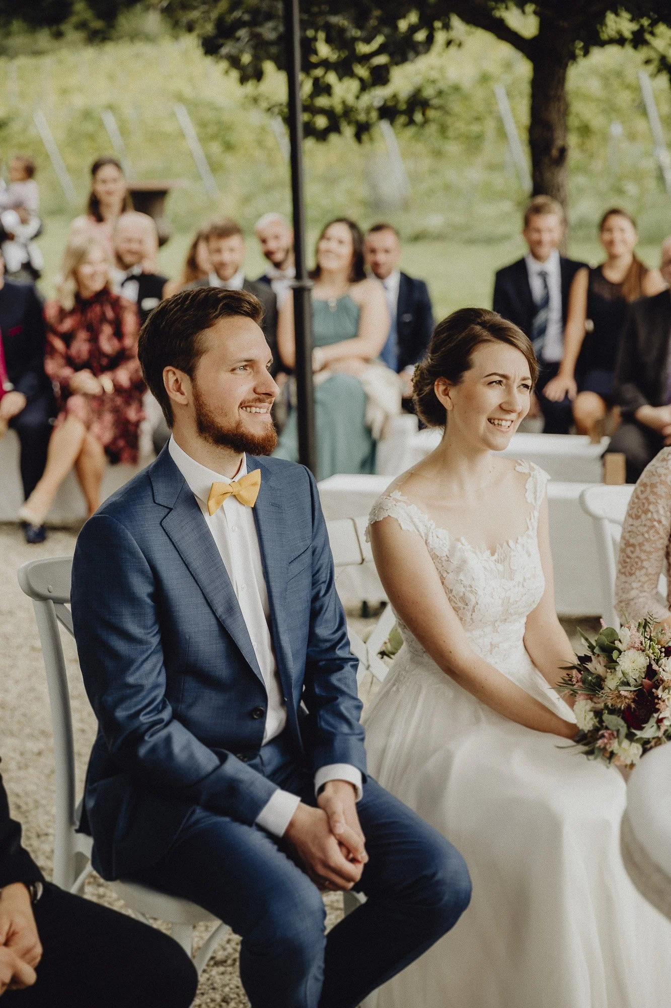 Hochzeit im Freien mit lachendem Brautpaar, sitzend auf weißen Stühlen, umgeben von Gästen, bei sonnigem Wetter unter Bäumen.