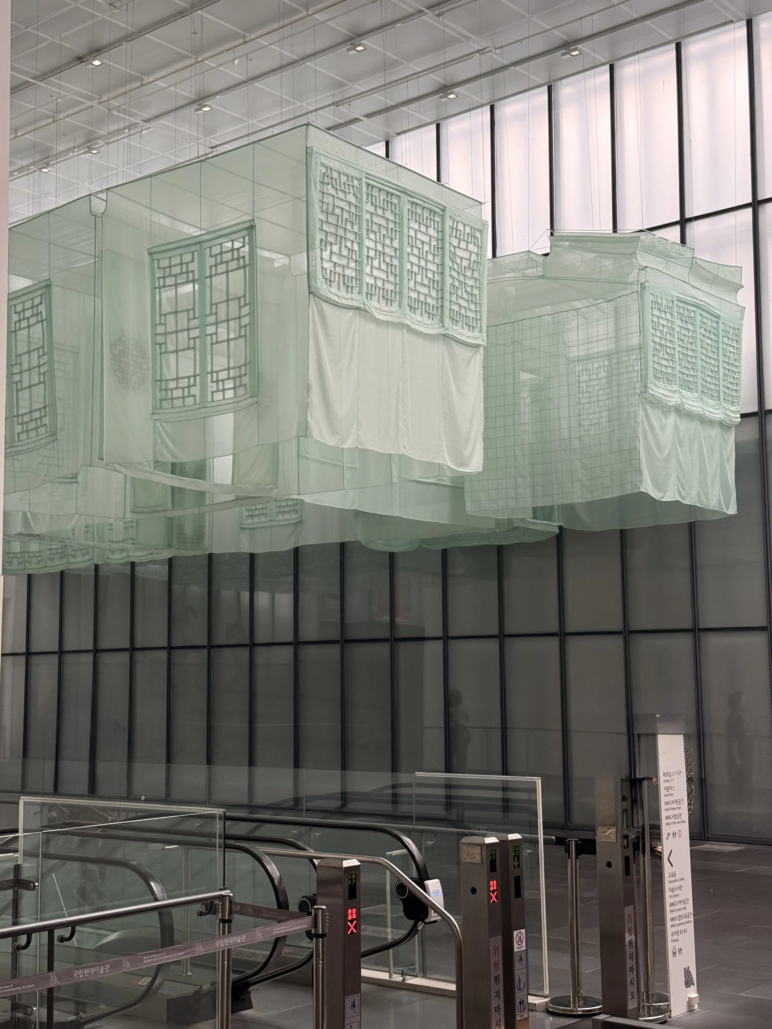 A pale green translucent fabric reconstruction of traditional Korean architecture by Do Ho Suh, suspended above the entrance area at MMCA Seoul.