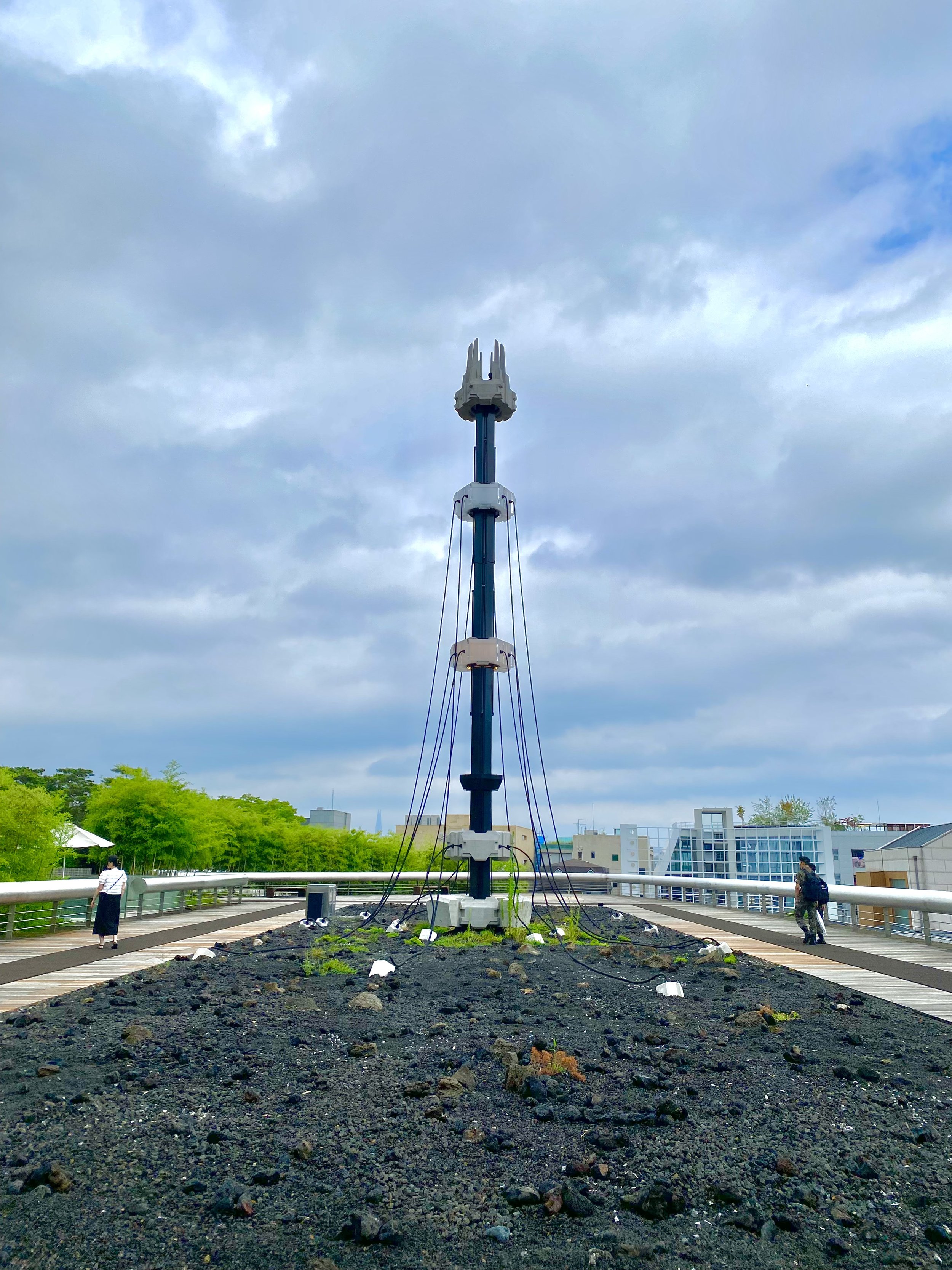 Tall pole with attached cables on a landscaped area with rocks, surrounded by a walkway and greenery under a cloudy sky.