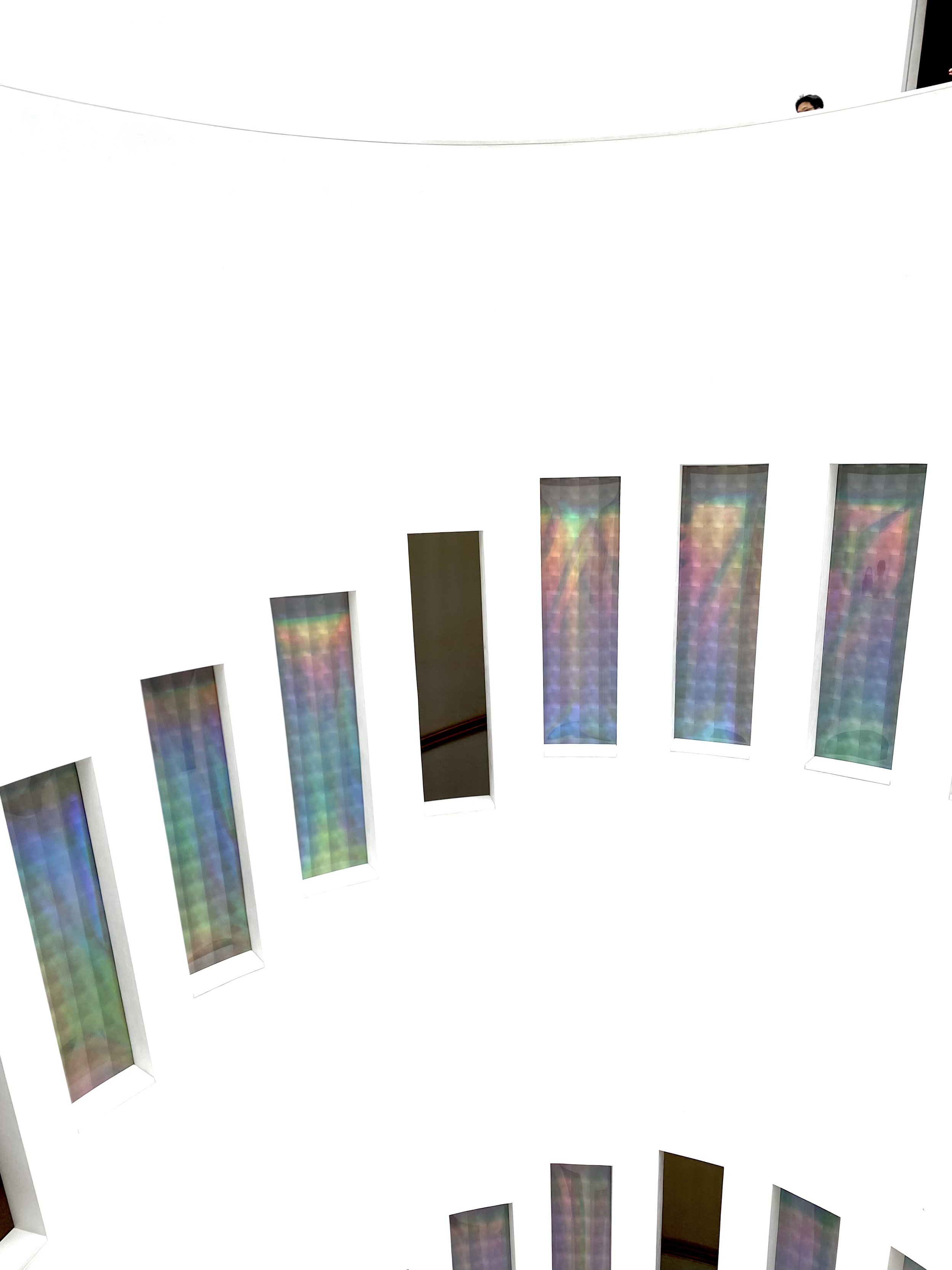 Curved white wall with vertical rainbow-colored windows and a small part of a person's head peeking over the wall.