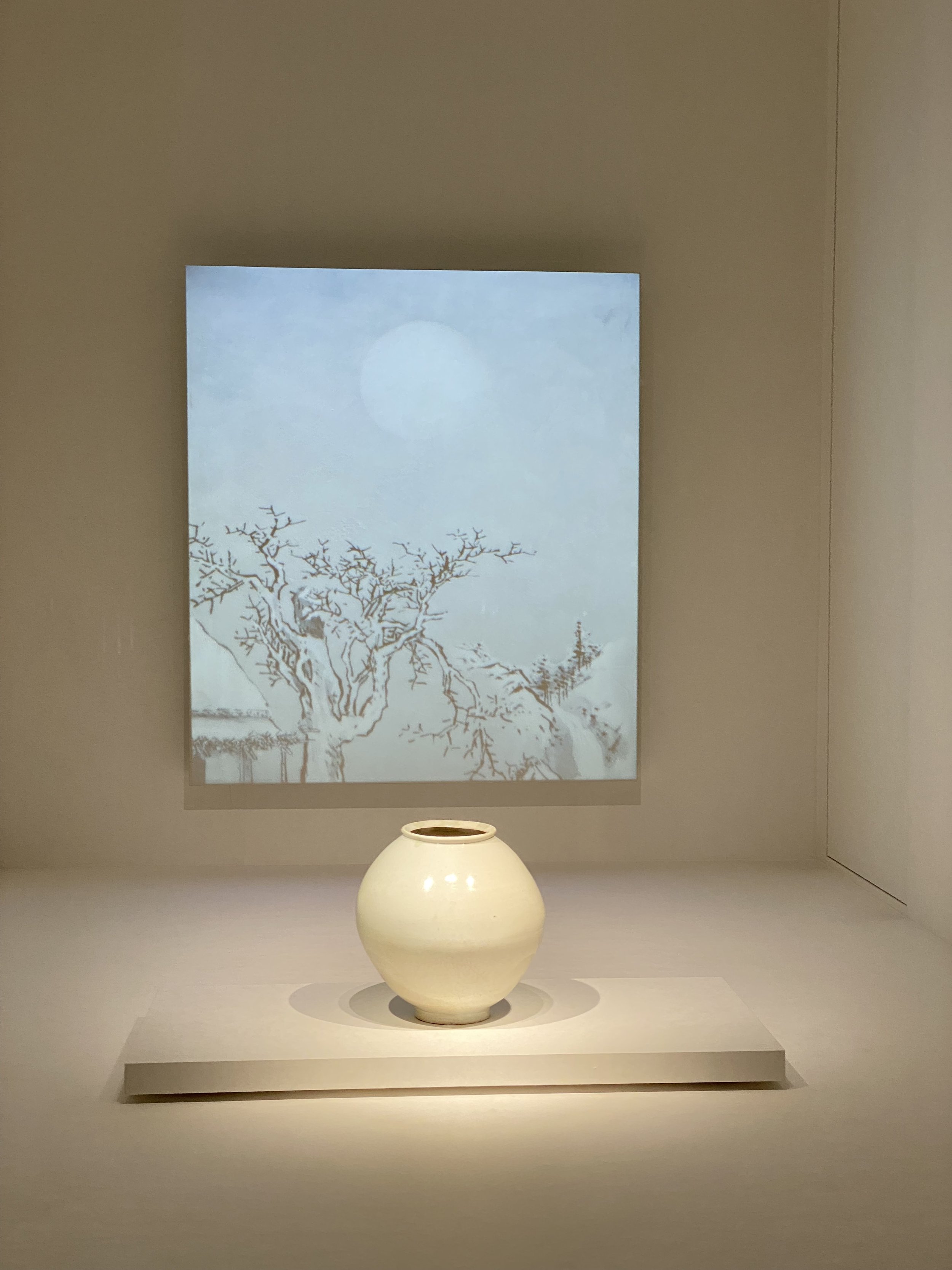 A white ceramic vase displayed on a pedestal in front of an artwork depicting a tree against a pale background.
