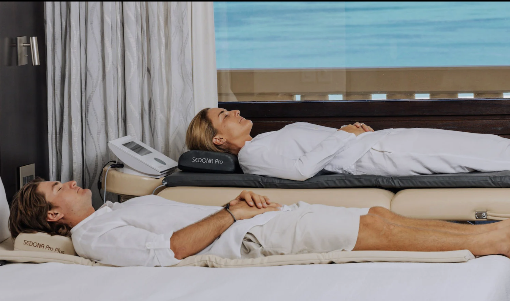 A man and a woman lying on beds side by side, relaxing with eyes closed, in a room with a large window showing a blue sky. The woman is on a sleep device labeled 'SEDONA Pro,' resting on a pillow that says 'SEDONA Pro Plus.'