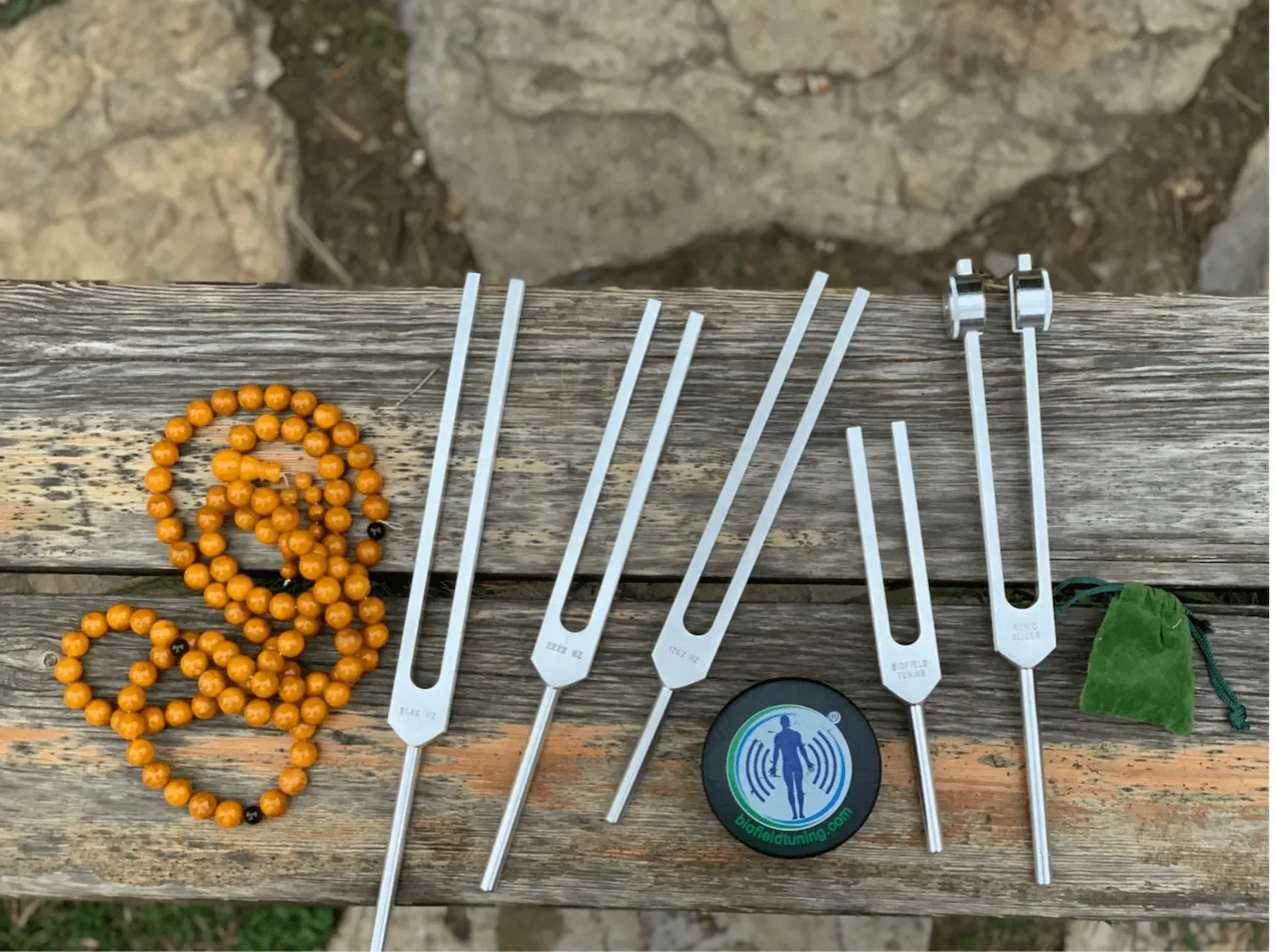 Various metal detectors with different probe sizes, an orange beaded necklace, a green drawstring pouch, and a round sticker with a human figure and the words 'biofieldduning.com,' all arranged on a wooden surface.