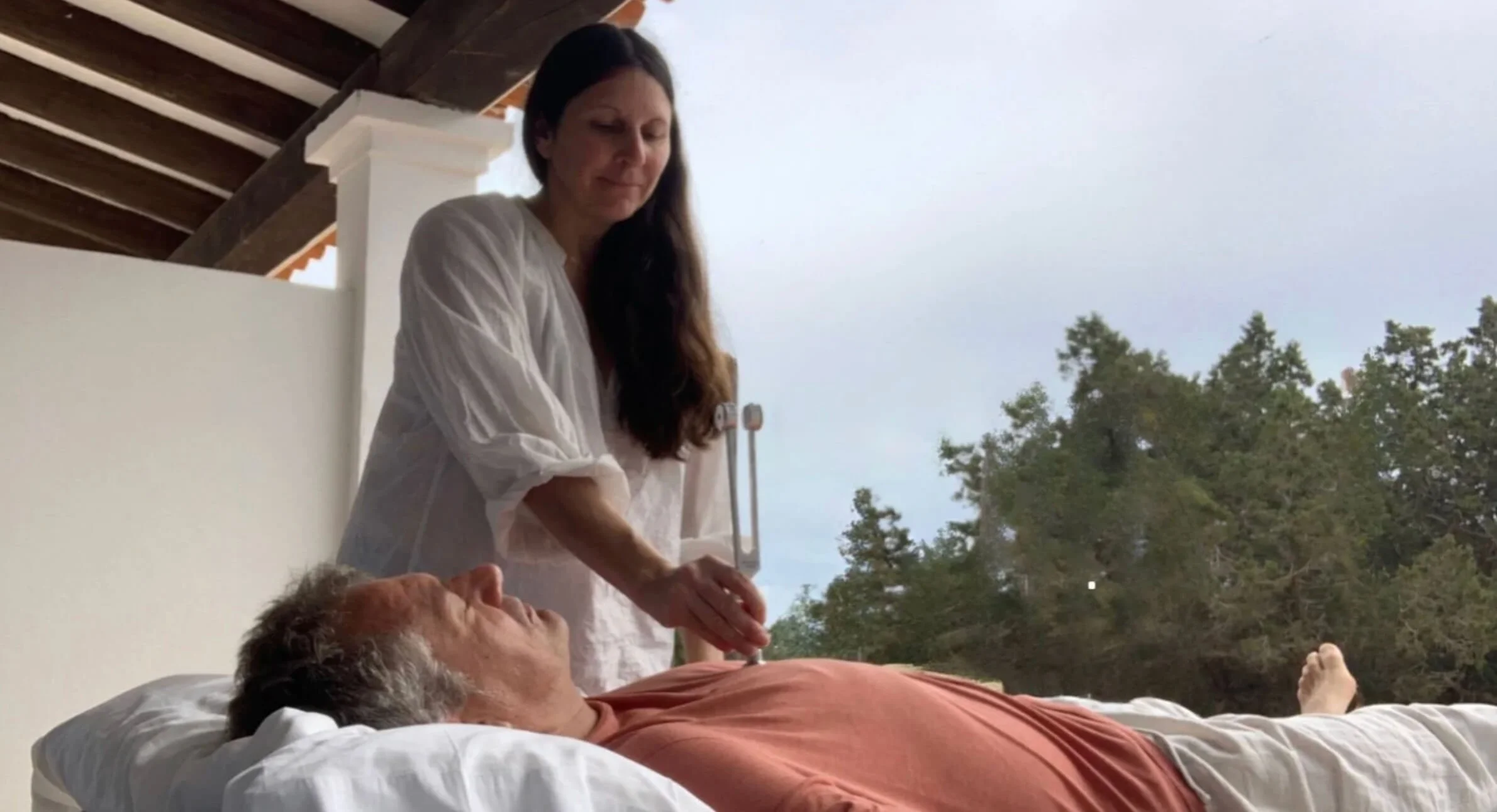 A woman dressed in white tending to an elderly man lying on a bed outside, with trees and a cloudy sky in the background.