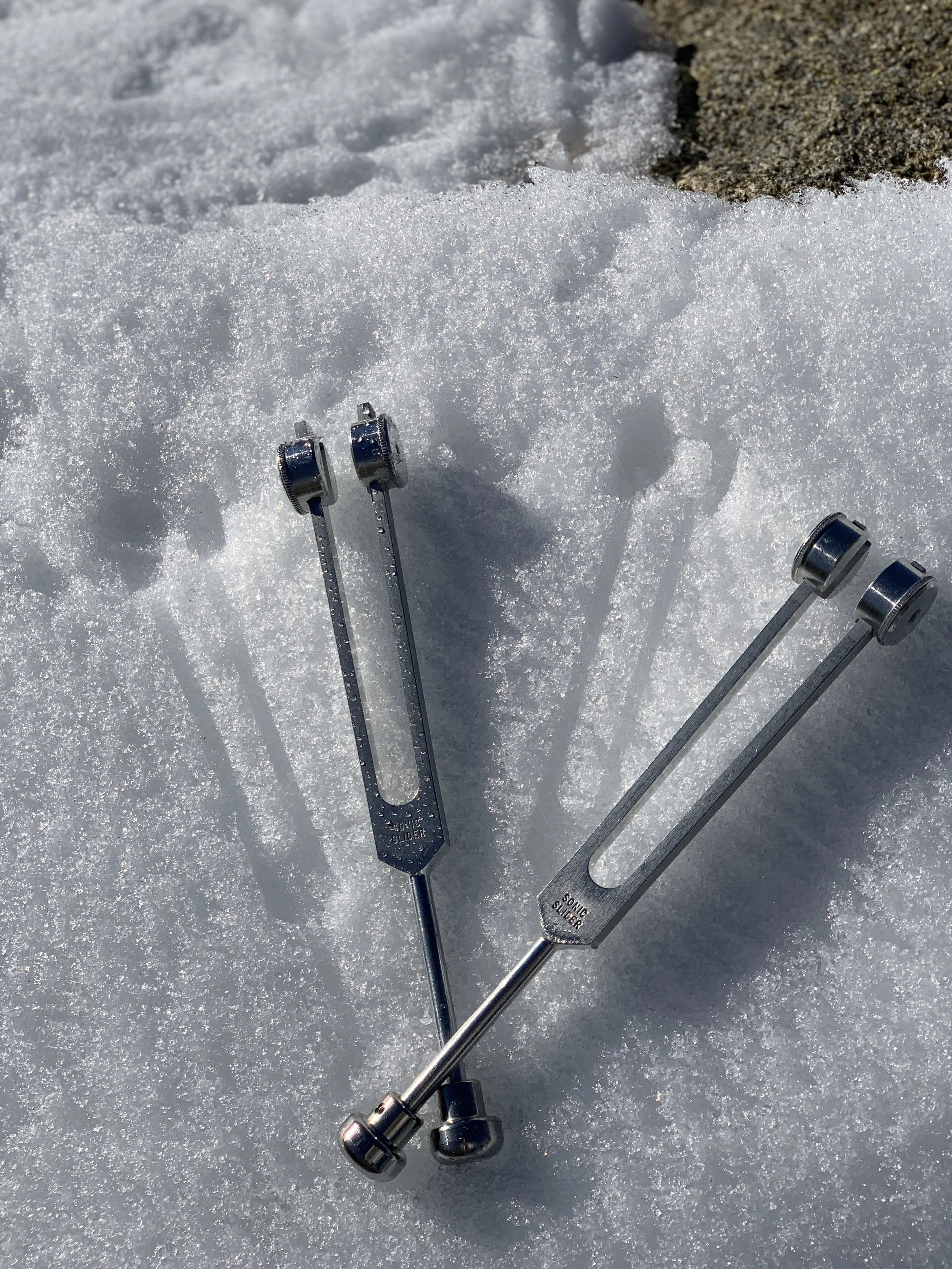 Two tuning forks placed on snow next to a rock, with their shadows cast on the snow.