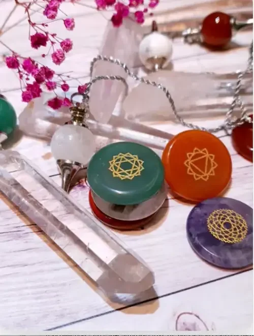 Colorful meditation tools and jewelry on a white wooden surface, including chakra stones and a clear crystal wand.