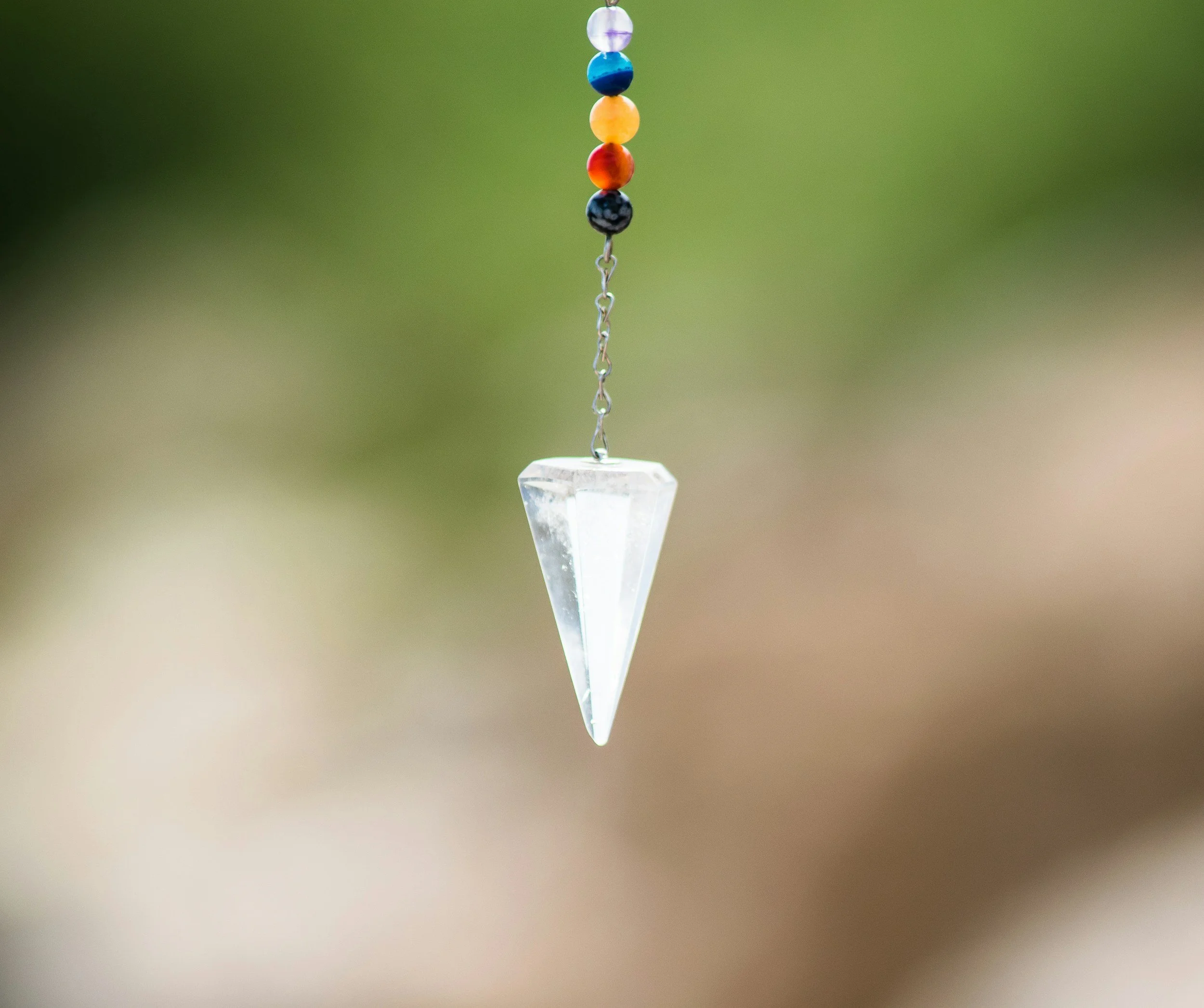 Hanging crystal prism pendant with colorful beads above, blurred green and beige background.