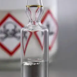 A glass vial with a metal cap, containing a clear liquid, with warning signs in the background indicating hazardous substances.