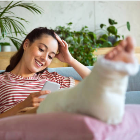 Young woman with a broken foot in a cast, smiling and looking at her phone while relaxing on a couch in a home setting.