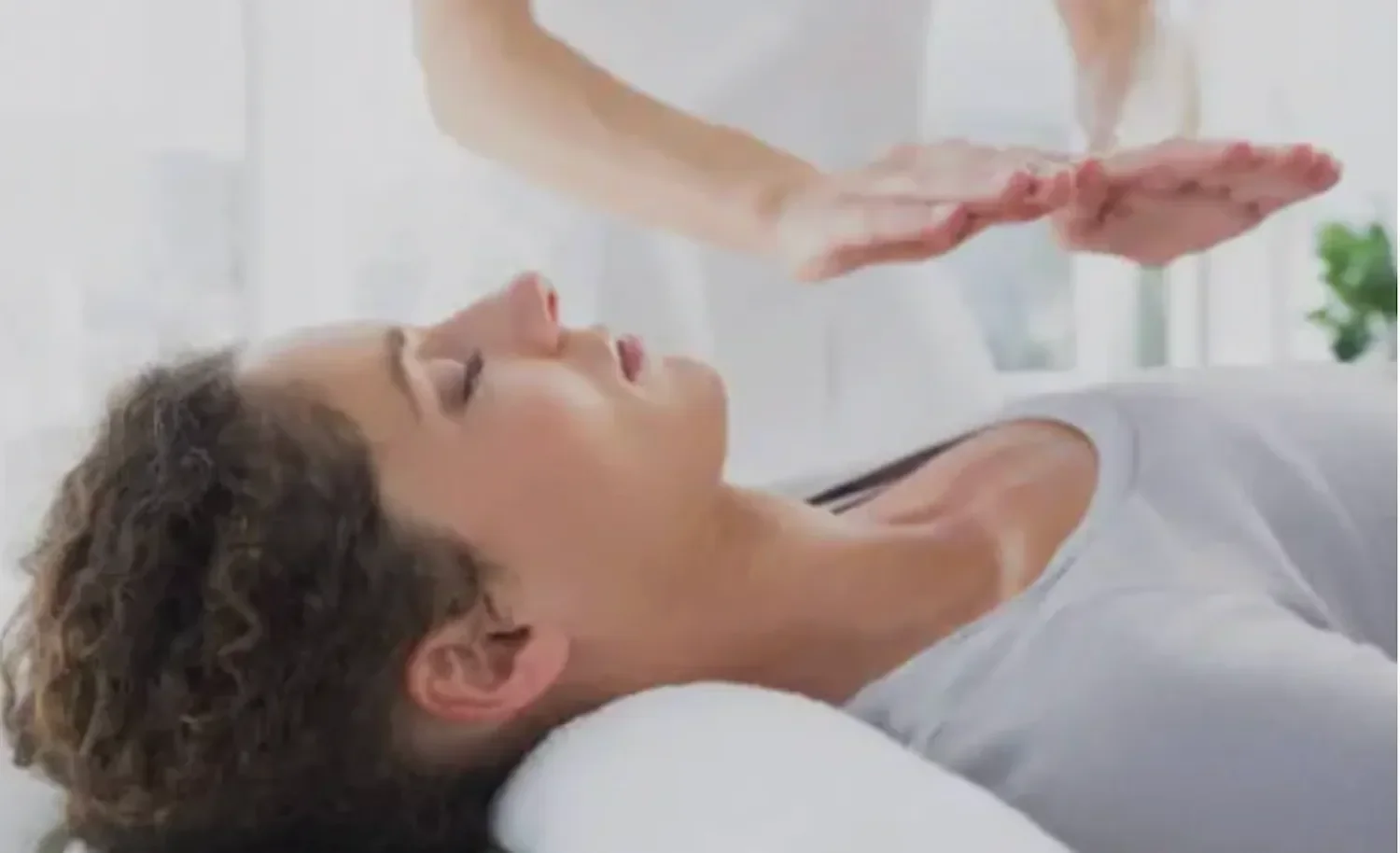 Person lying on a bed with their eyes closed, appearing to be receiving a massage or relaxation treatment from another person whose hands are visible above them.