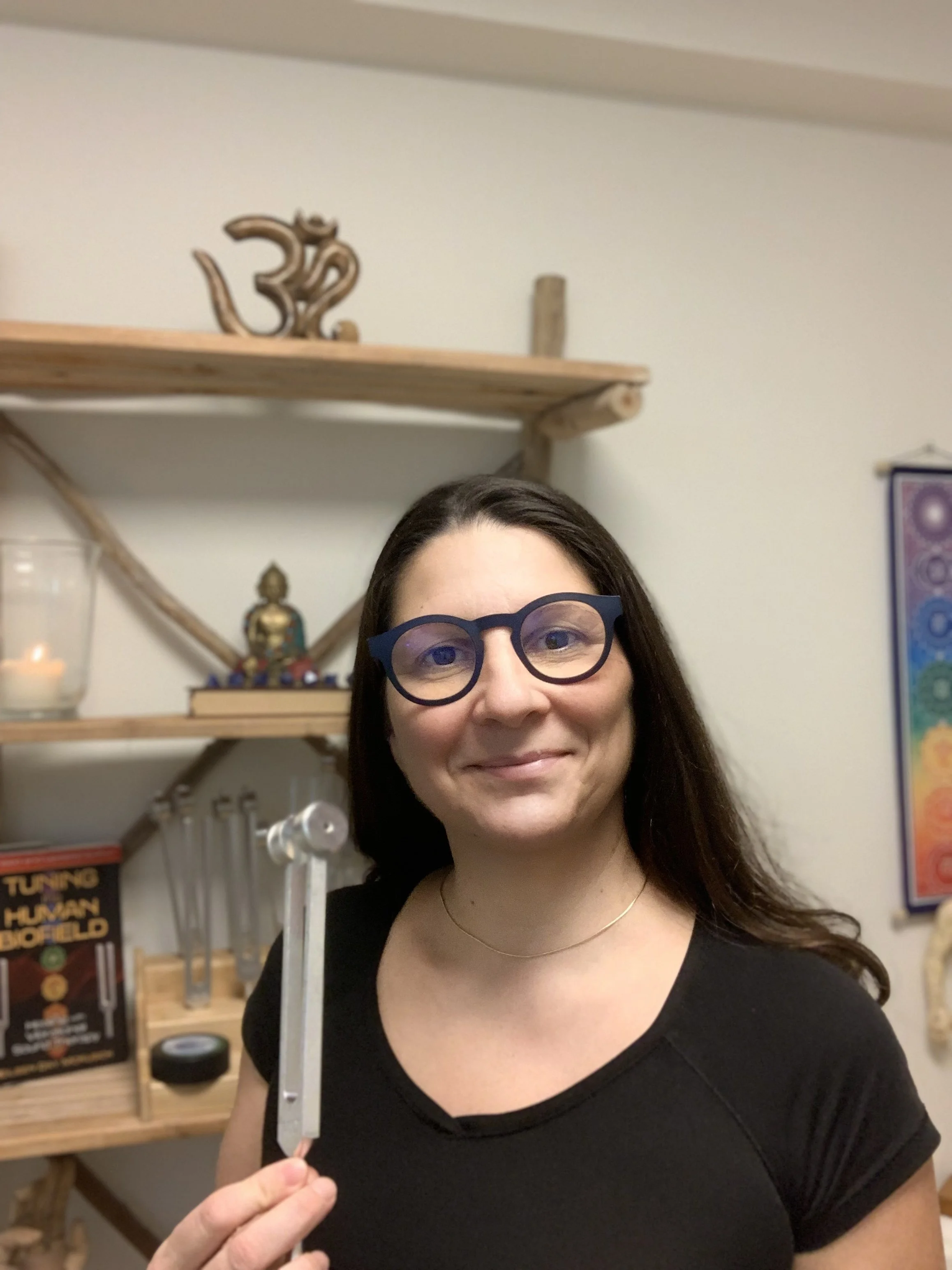 A woman wearing glasses and a black shirt, smiling, standing indoors with shelves and spiritual items behind her.