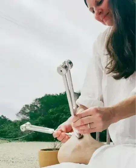 A woman in white clothes is performing a medical or research procedure on her abdomen outdoors, using a metal instrument with cylindrical parts, with trees and a cloudy sky in the background.