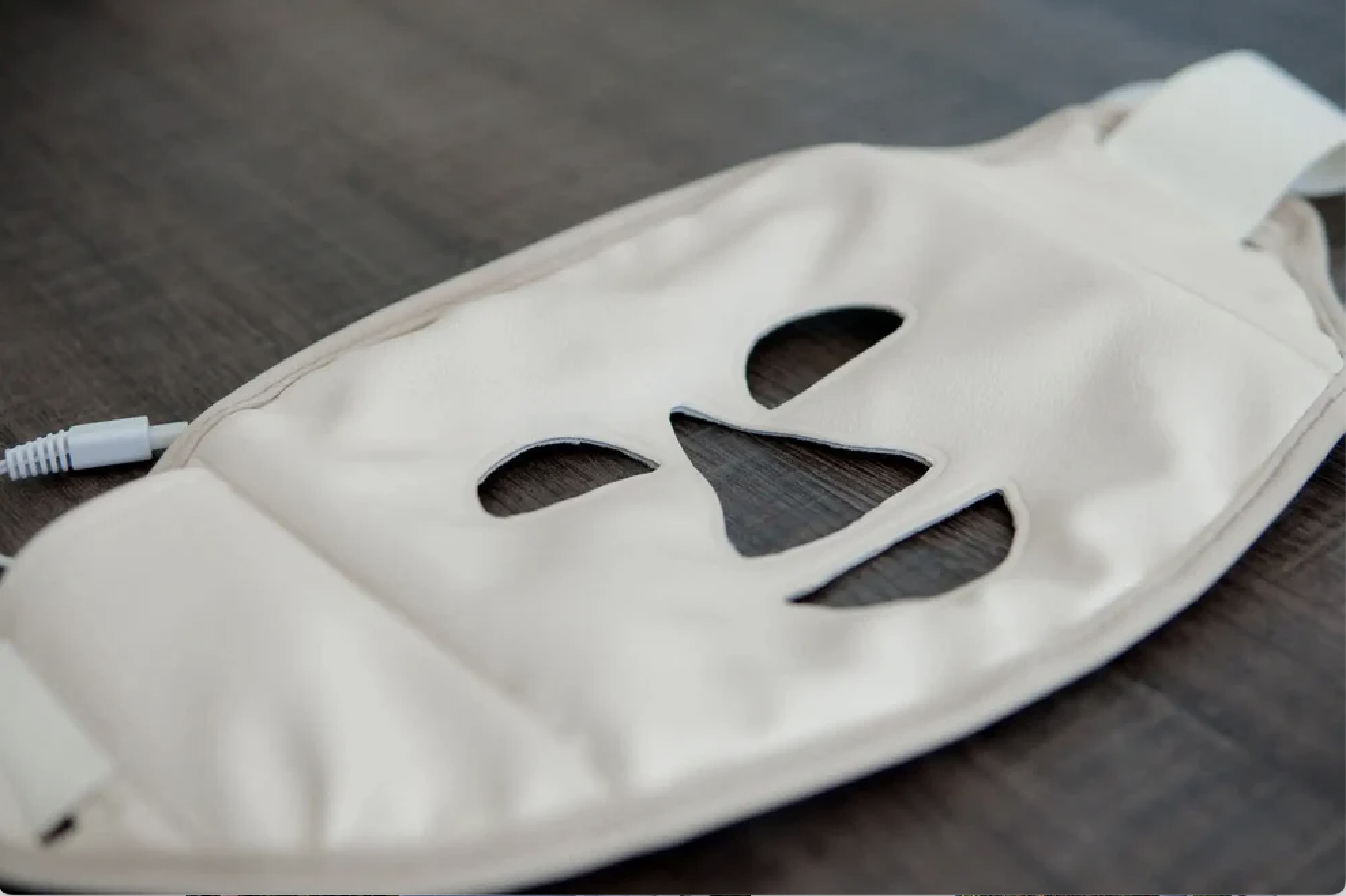 A white facial treatment mask with cutouts for eyes, nose, and mouth, lying on a dark wooden surface.