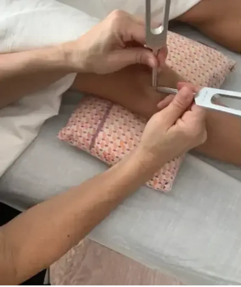 Medical procedure involving the insertion of a needle into a patient's leg, with a healthcare worker holding surgical tools and the patient lying on a padded surface.
