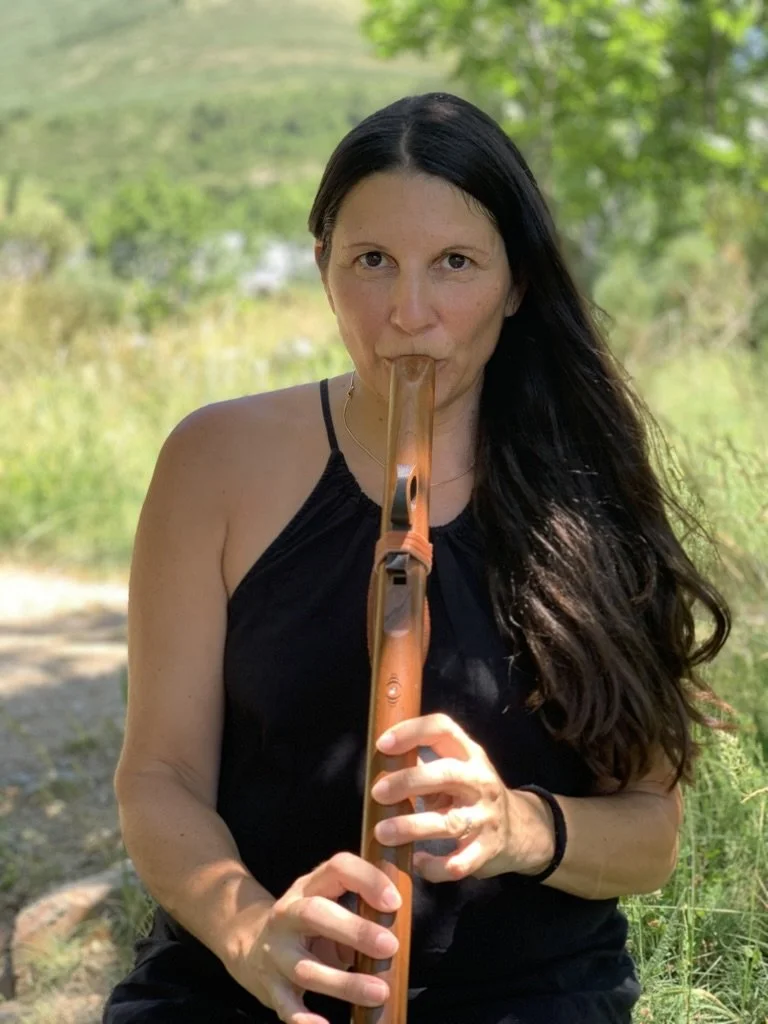 Woman playing a wooden flute outdoors in a green, natural setting.