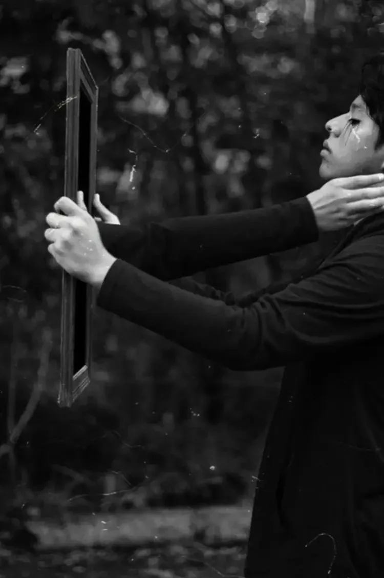 A person holding a mirror with their left hand, while their right hand touches their neck. The person is wearing a black long-sleeve shirt and has tears on their face. The background is blurred, with trees and foliage visible.