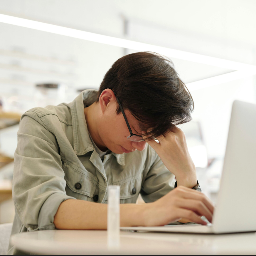 Young man with glasses resting his head on his hand, sitting at a desk with a laptop in front of him, appearing stressed or tired.