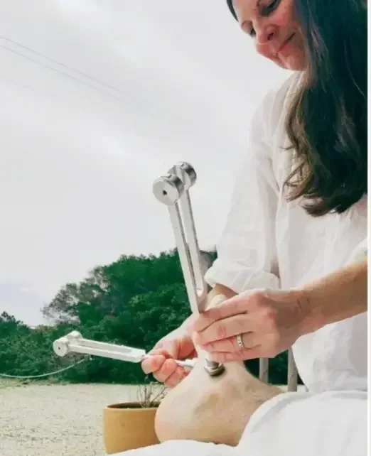 A woman in white working on a pottery piece outdoors, using large metal tools for shaping clay.