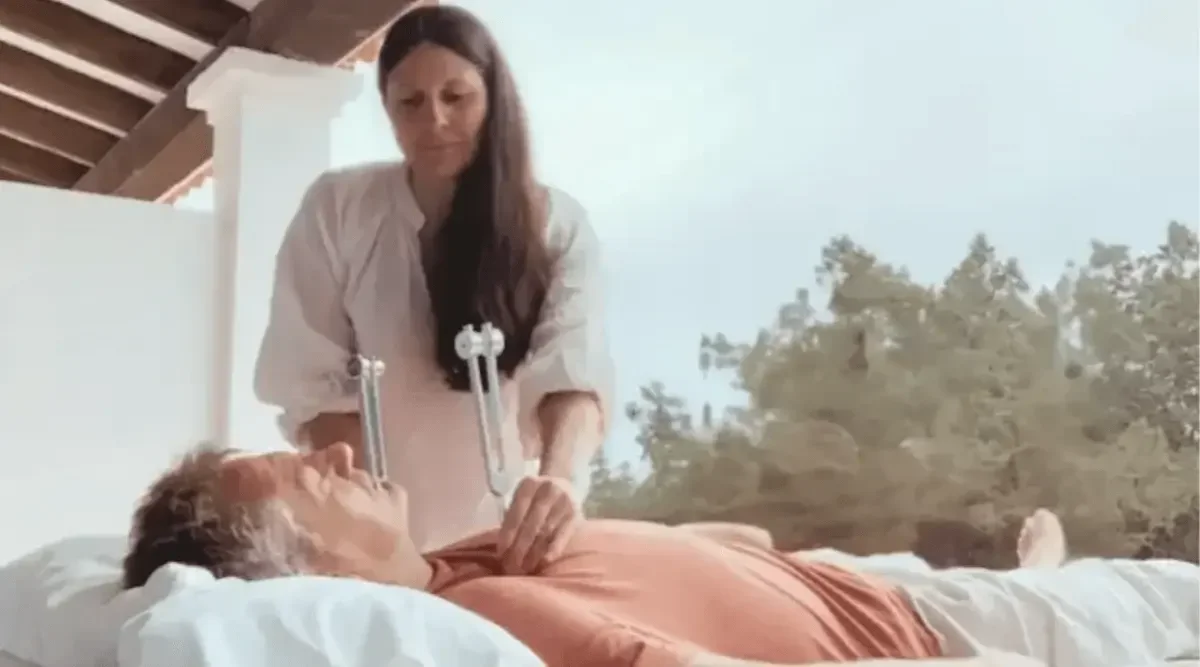 A woman in white medical attire tending to an elderly man lying in bed, with trees and outdoor scenery visible through a window.