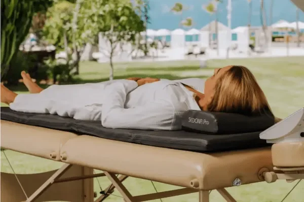 A woman lying on a massage table outdoors, with trees, a fence, and tents visible in the background.