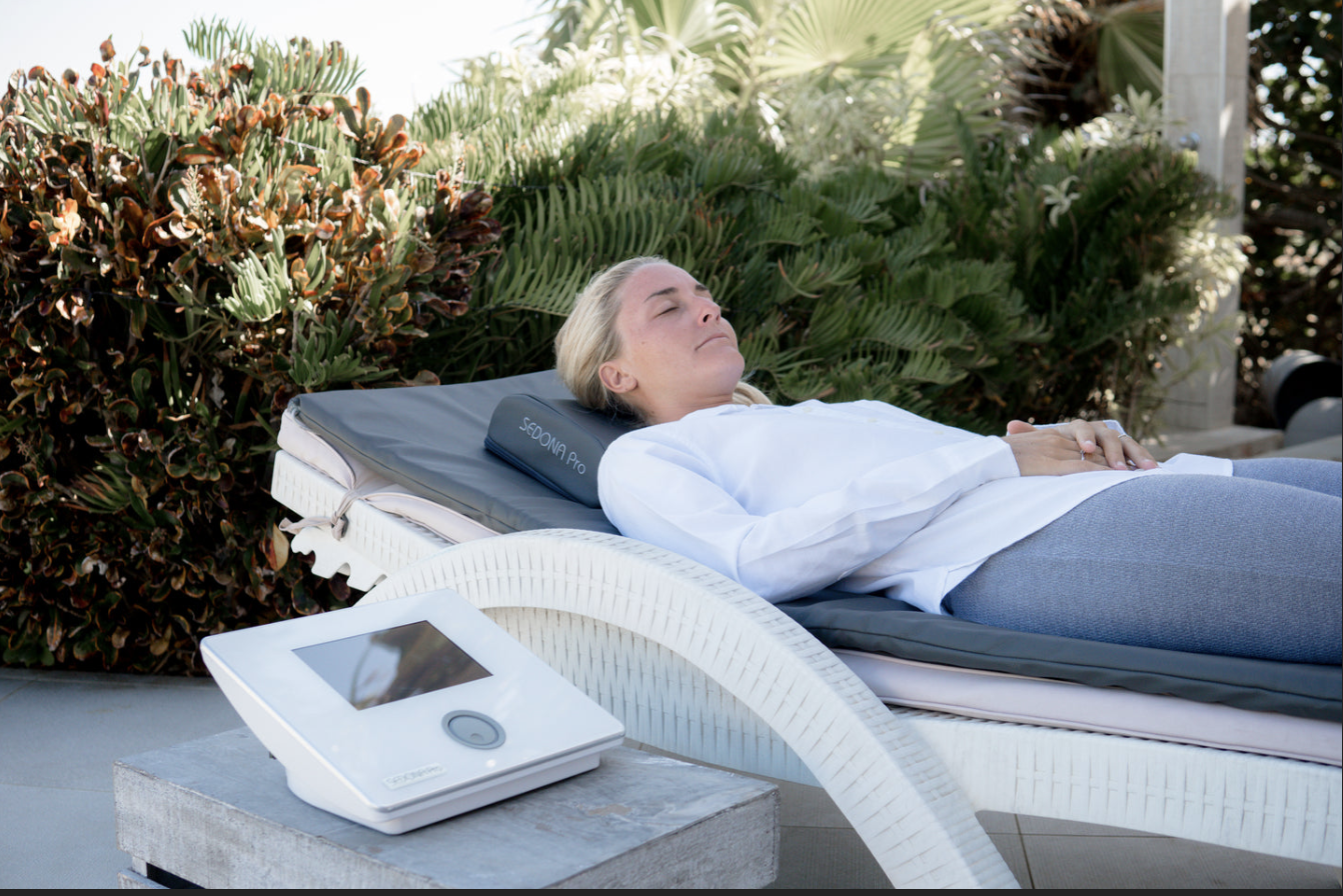 A woman is lying on a lounge chair outdoors, resting with her eyes closed, on a sunny day. There is a nearby device on a small table, and lush green foliage surrounds her.