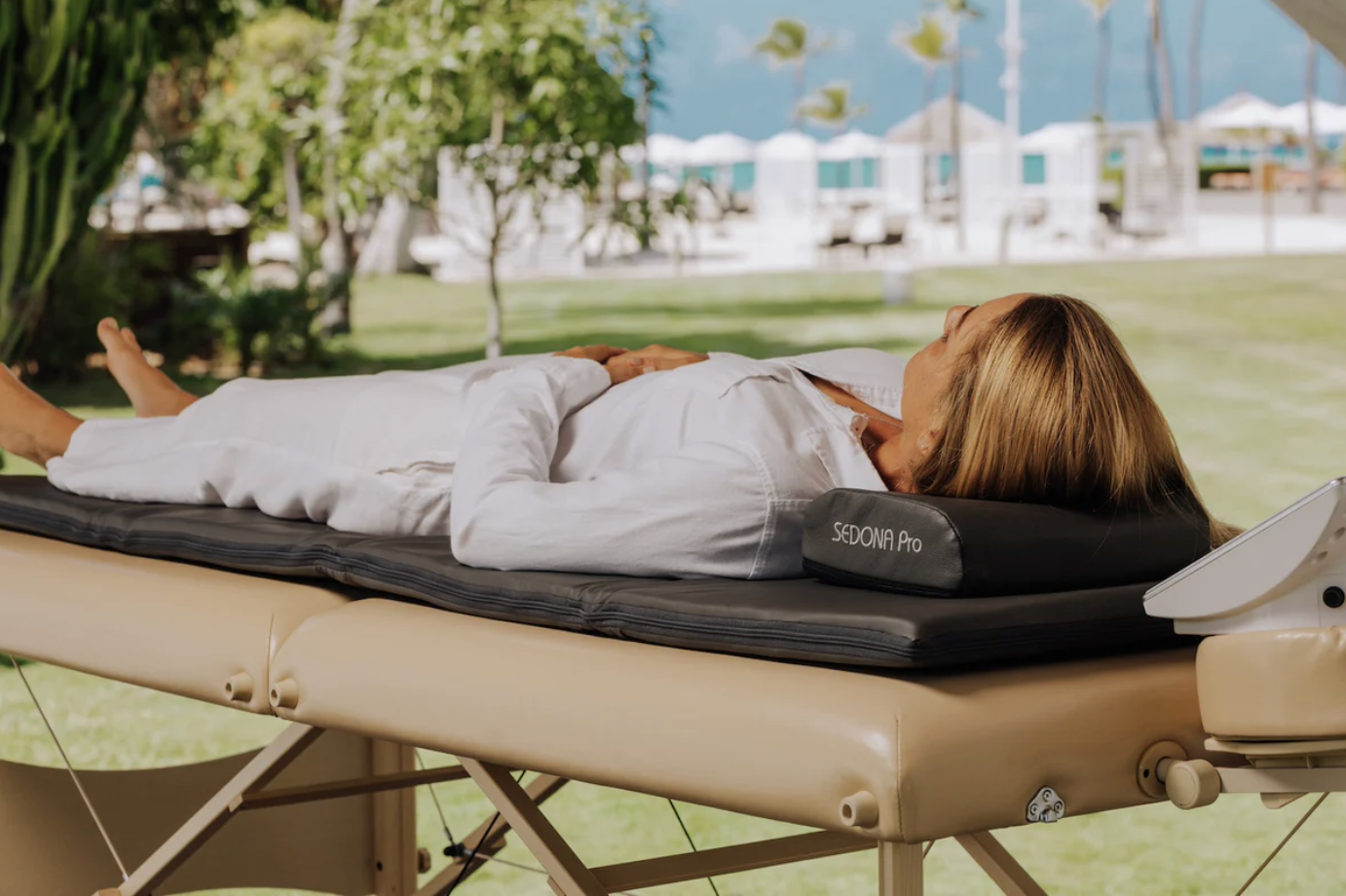 A woman lies on a massage table outdoors, with her head resting on a black pillows labeled 'Sedona Pro'. She appears relaxed, with her arms resting on her stomach. The background displays green trees, a swimming pool, and beach chairs under umbrellas, suggesting a resort setting.
