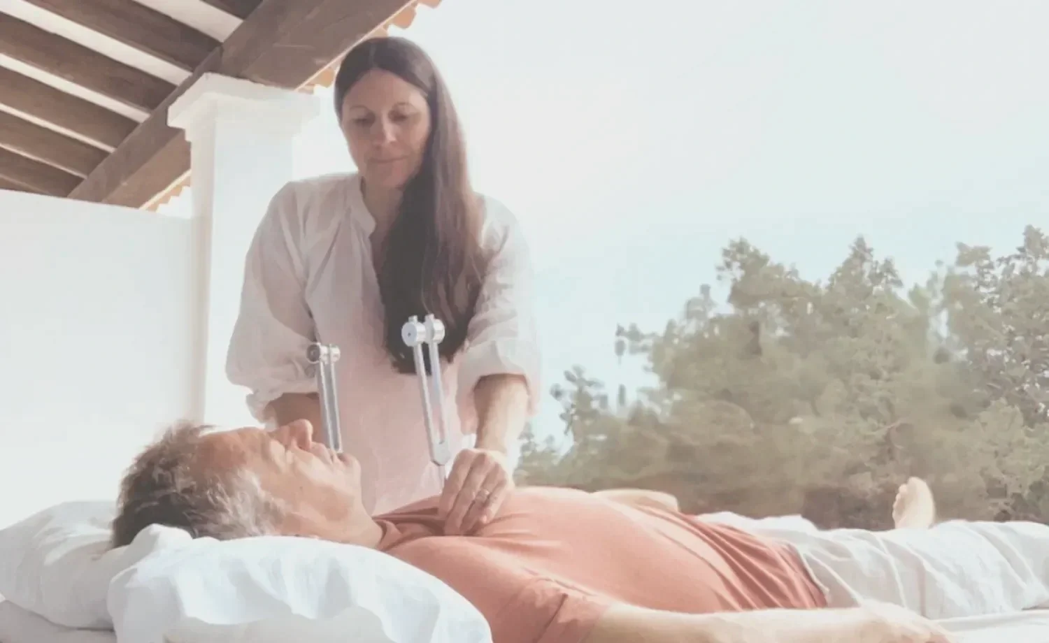 A woman tending to an elderly man lying in bed outdoors, with trees and a blue sky in the background.