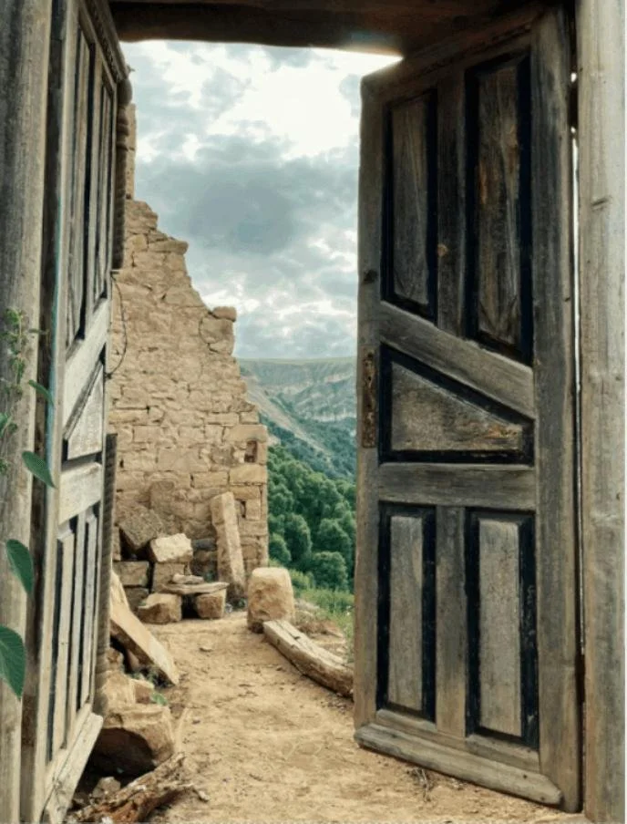 An old rustic wooden door open to reveal a view of green hills and cloudy sky beyond, with ruins of stone walls inside.