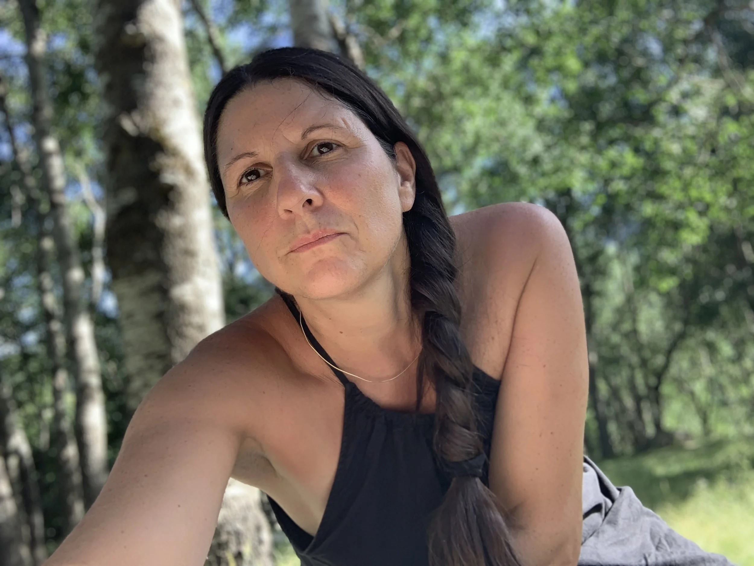 A woman with long dark hair in a braid, wearing a black top, outdoors in a wooded area with green trees in the background, looking at the camera.