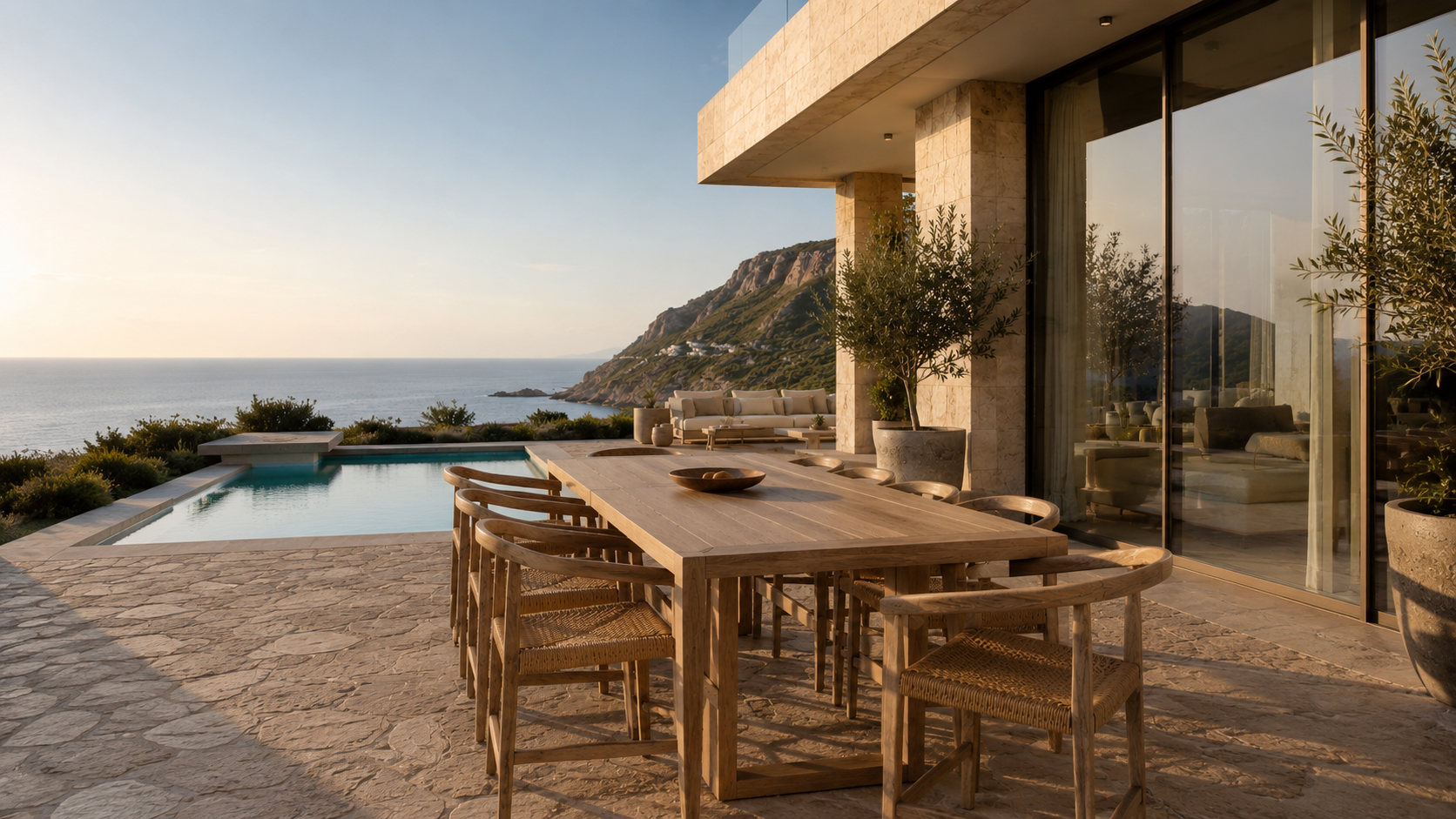 A modern outdoor patio with a wooden dining table and chairs, overlooking a swimming pool and ocean view, with a mountainous coastline in the background at sunset.