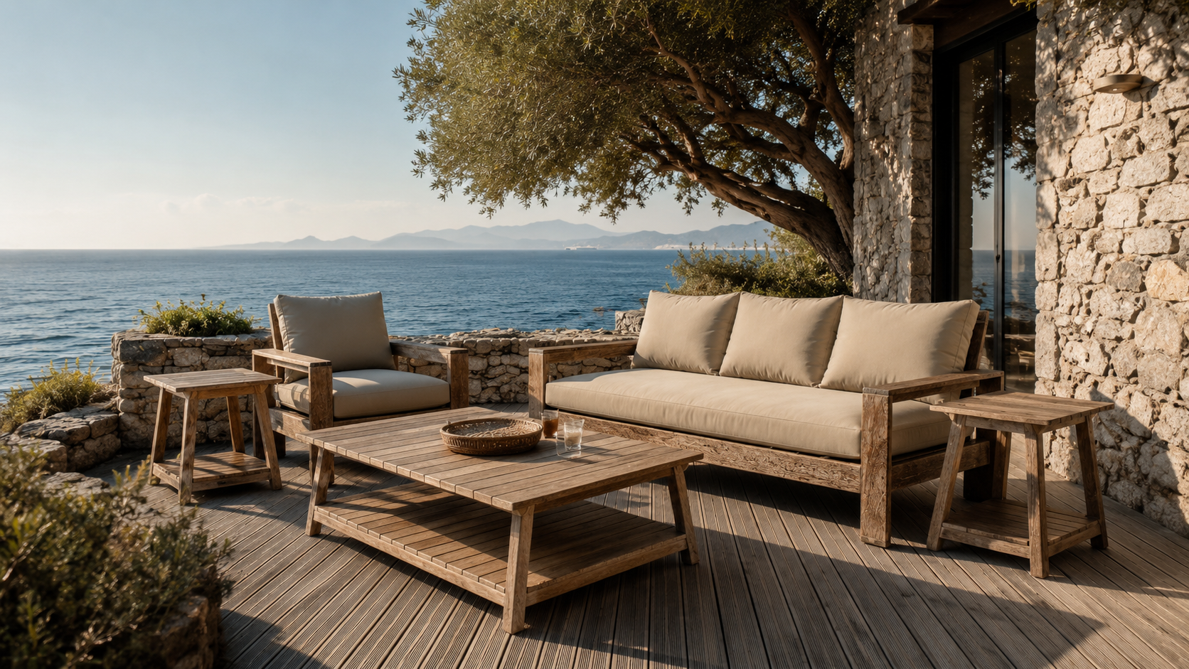 Outdoor patio with wooden furniture, cushioned chairs and sofa, overlooking the ocean with distant mountains and a tree providing shade.