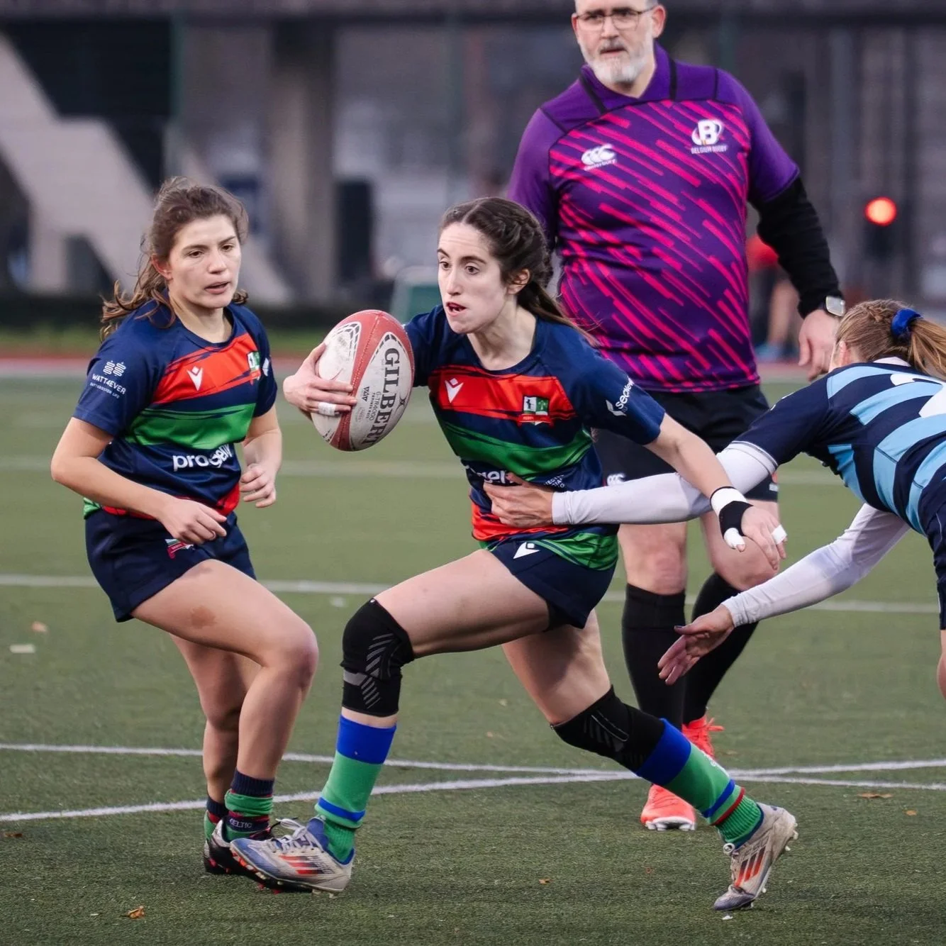 Vrouwen spelen rugby op een veld, één rent met de bal in een groen en blauw shirt, anderen in de achtervolging.