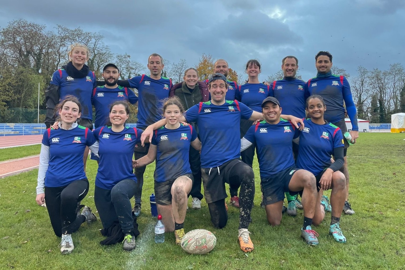 Groupe de joueurs de rugby en maillots bleus posant sur un terrain, avec des hommes et des femmes souriant et s'agenouillant ensemble, tenant un ballon de rugby.