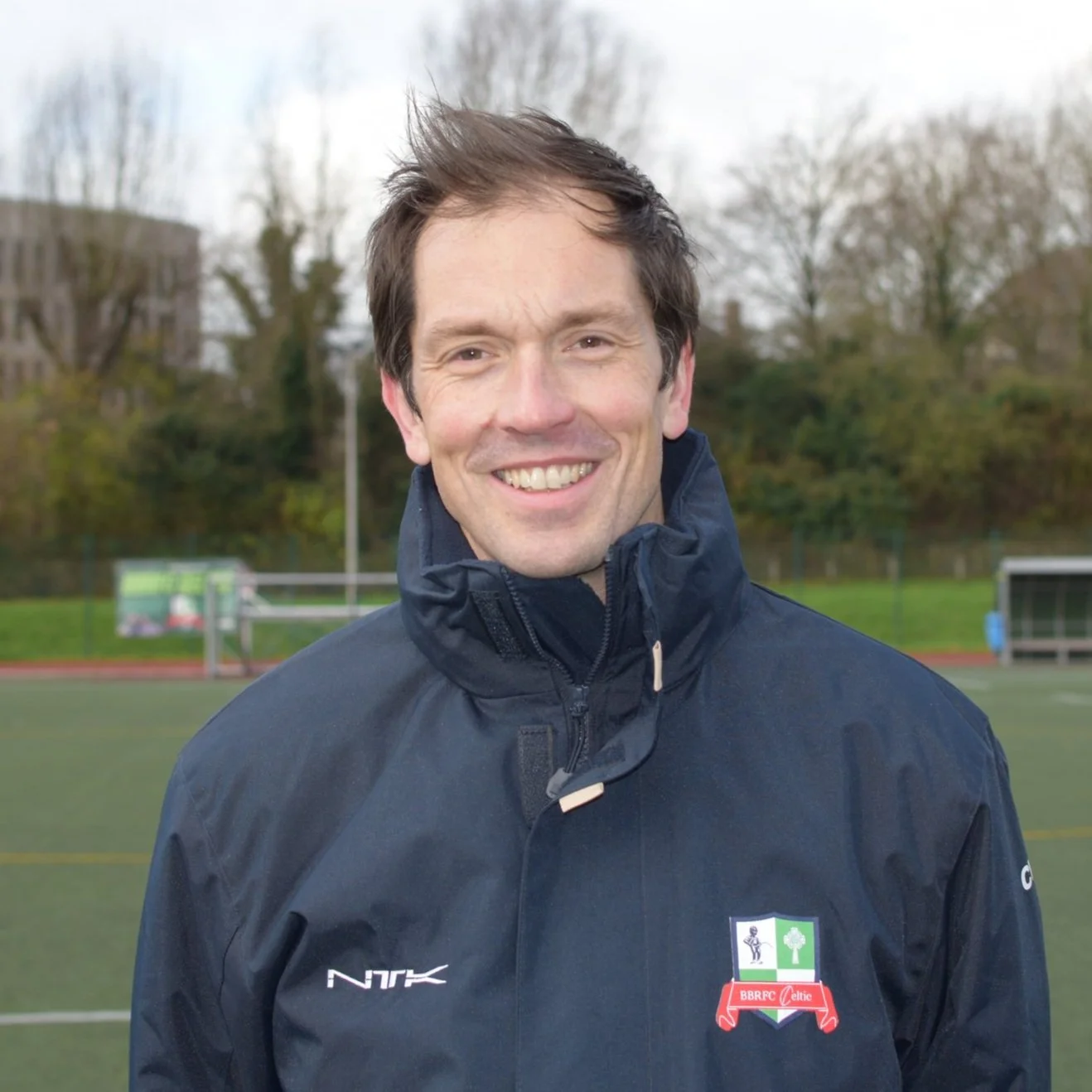 Persoon die buiten glimlacht en een donker jack draagt met een sportlogo