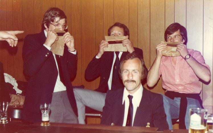 Four men sitting in a bar with three holding paper bags to their mouths, another man sitting with a mustache, wood-paneled background, and drinks on the table.