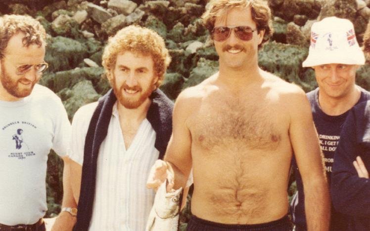 Four men standing by rocky shoreline, one shirtless holding a fish, wearing sunglasses.