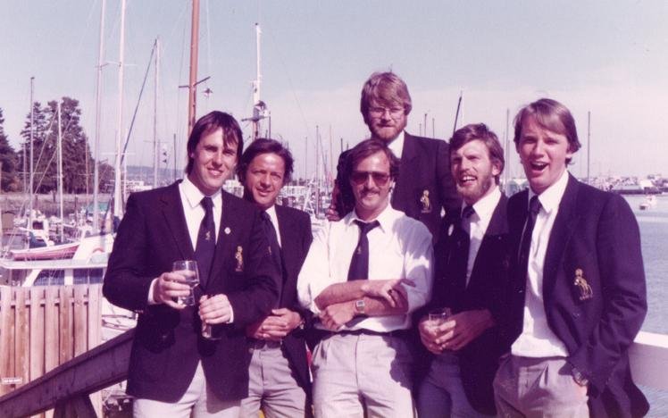 Group of men in blazers standing by a marina with sailboats