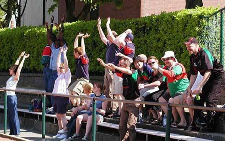 Group of people cheering at an outdoor event, sitting on bleachers, with some holding drinks and others raising arms.