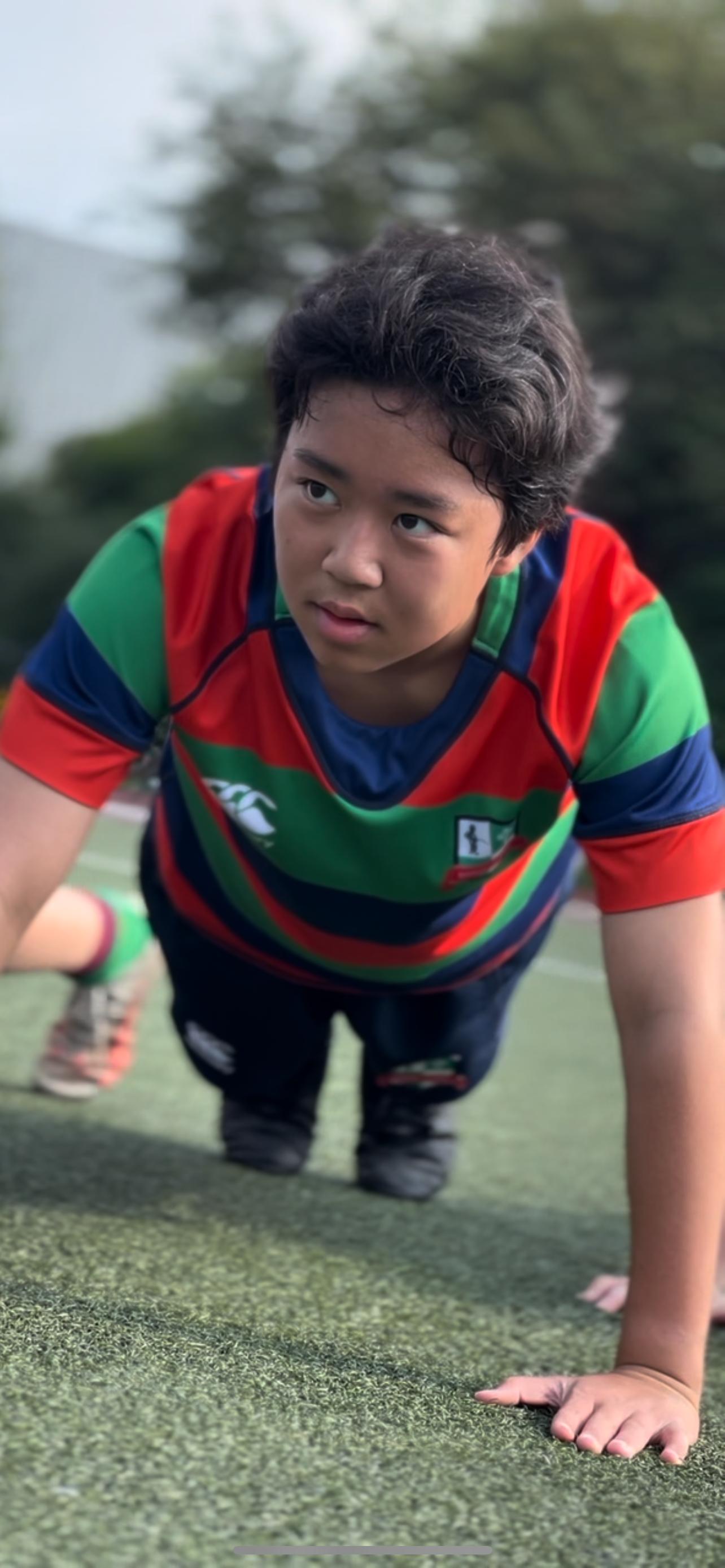 A young individual wearing a colorful striped sports jersey is on a grassy field, positioned in a crawling stance and looking forward, suggesting focus and concentration in an outdoor athletic setting.